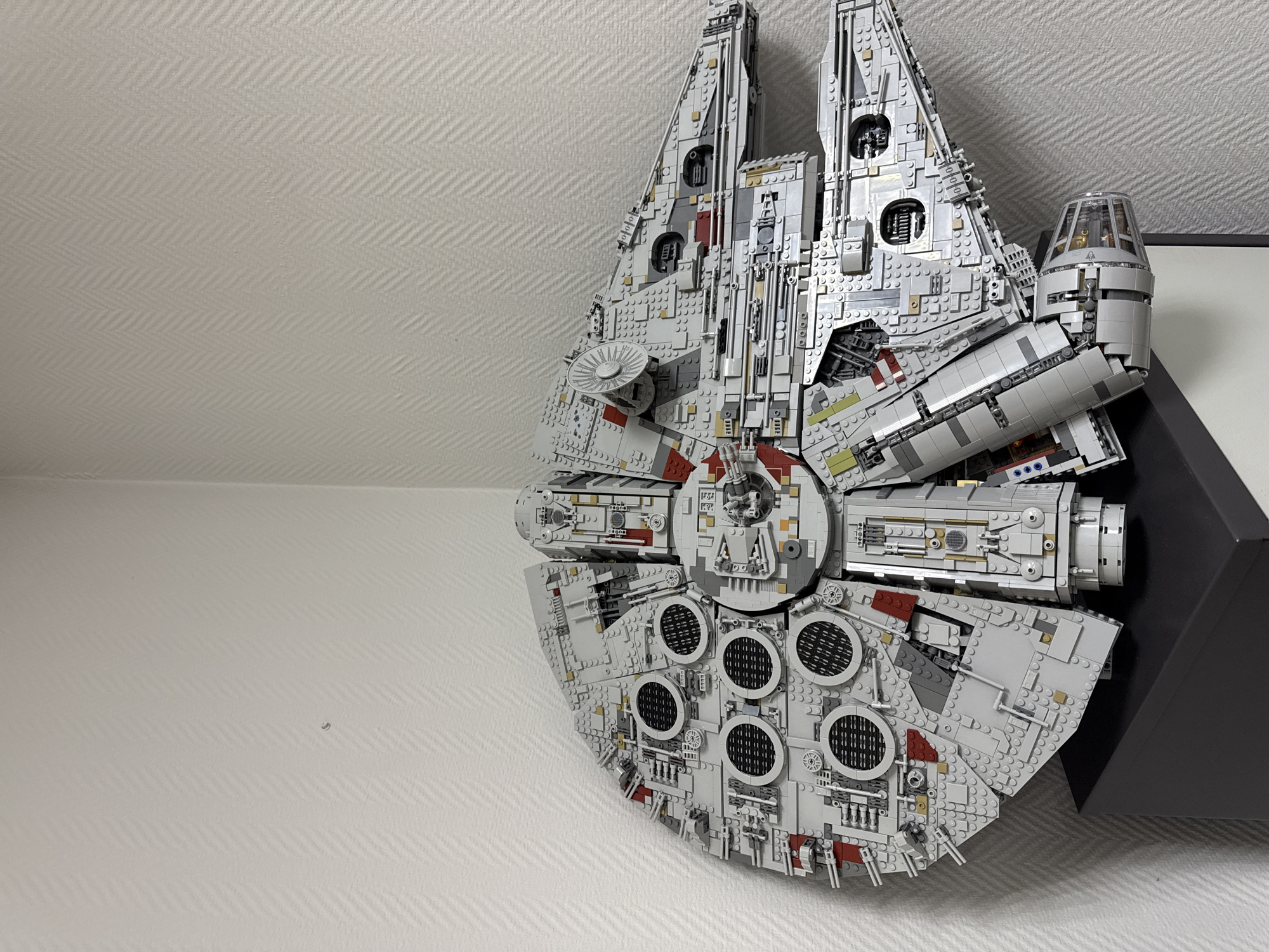 A grey Lego Millennium Falcon model is mounted on a white wall with a subtle zigzag pattern. Part of a black frame or shelf supports the model on the right side of the image. The wall appears to be indoors, possibly in a home or office setting. The scene does not contain any information to determine the city of Krasnoyarsk, Russia.