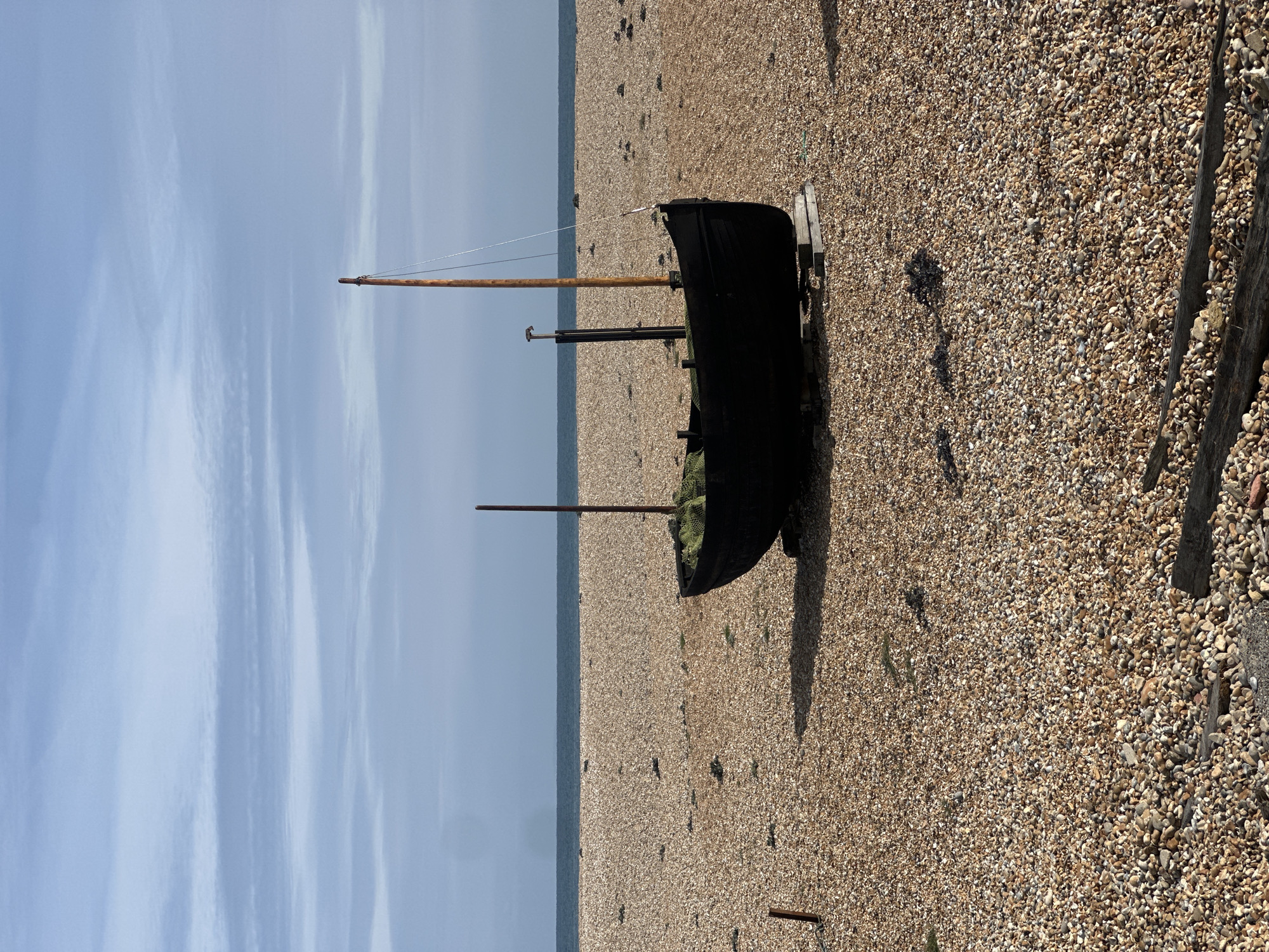 The image shows an old, dark-colored boat resting on a pebble beach. The boat has masts and is supported by wooden blocks. The beach extends toward the sea and the sky is blue with white clouds. This scene likely takes place in the Folkestone and Hythe District, United Kingdom.