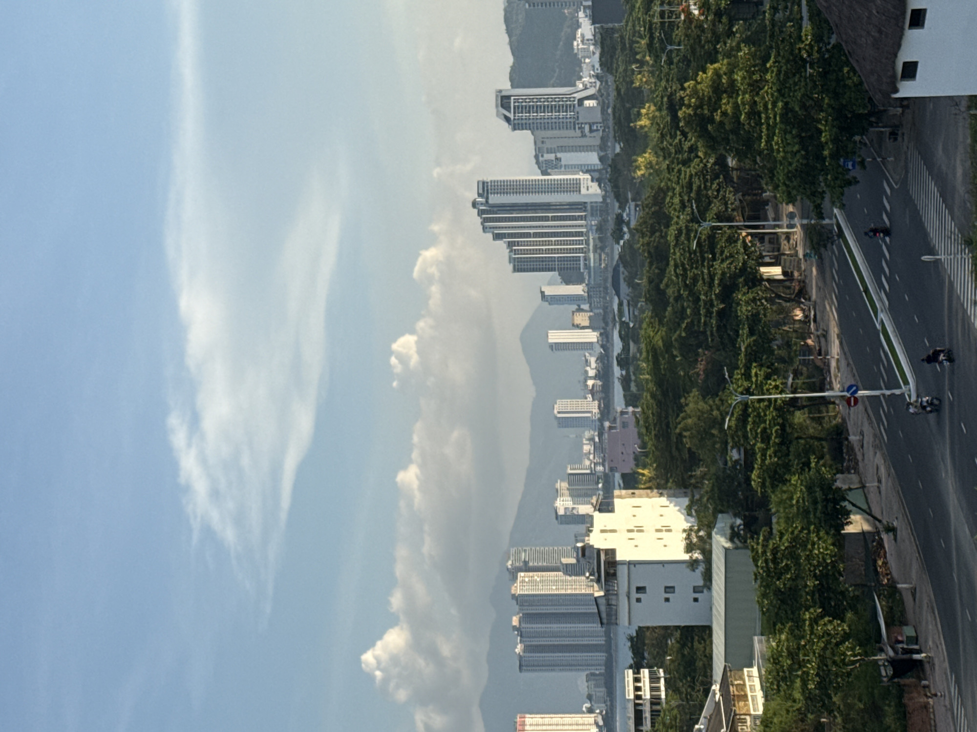 The image captures a panoramic view of a coastal city, likely Nha Trang, Vietnam, characterized by a vibrant urban landscape juxtaposed with lush greenery and a mountainous backdrop under a bright, partly cloudy sky. In the foreground, a street with clear lane markings and a pedestrian crossing is visible, with a few motorcycles and a single scooter in motion. A street lamp stands on the right side of the road, accompanied by traffic signs. Adjacent to the road, a cluster of trees with dense foliage line the area, interspersed with some low-rise buildings and structures, including a white building with visible windows and a thatched-roof structure on the far right. Further into the mid-ground, more trees dominate the landscape, concealing parts of the city. A body of water, possibly the sea, separates the foreground and mid-ground from the more distant cityscape. The background is dominated by a skyline of numerous tall buildings and high-rises, showcasing modern architecture. Beyond the city, a range of mountains stretches across the horizon, their peaks partially obscured by clouds. The sky is a clear blue with scattered white clouds, some of which are large and puffy, suggesting fair weather. The lighting indicates it is likely late afternoon or early morning, with a soft, warm glow cast over the scene. The presence of high-rise buildings and the general urban layout strongly suggests a city like Nha Trang, a popular tourist destination in Vietnam known for its beaches and coastal development.