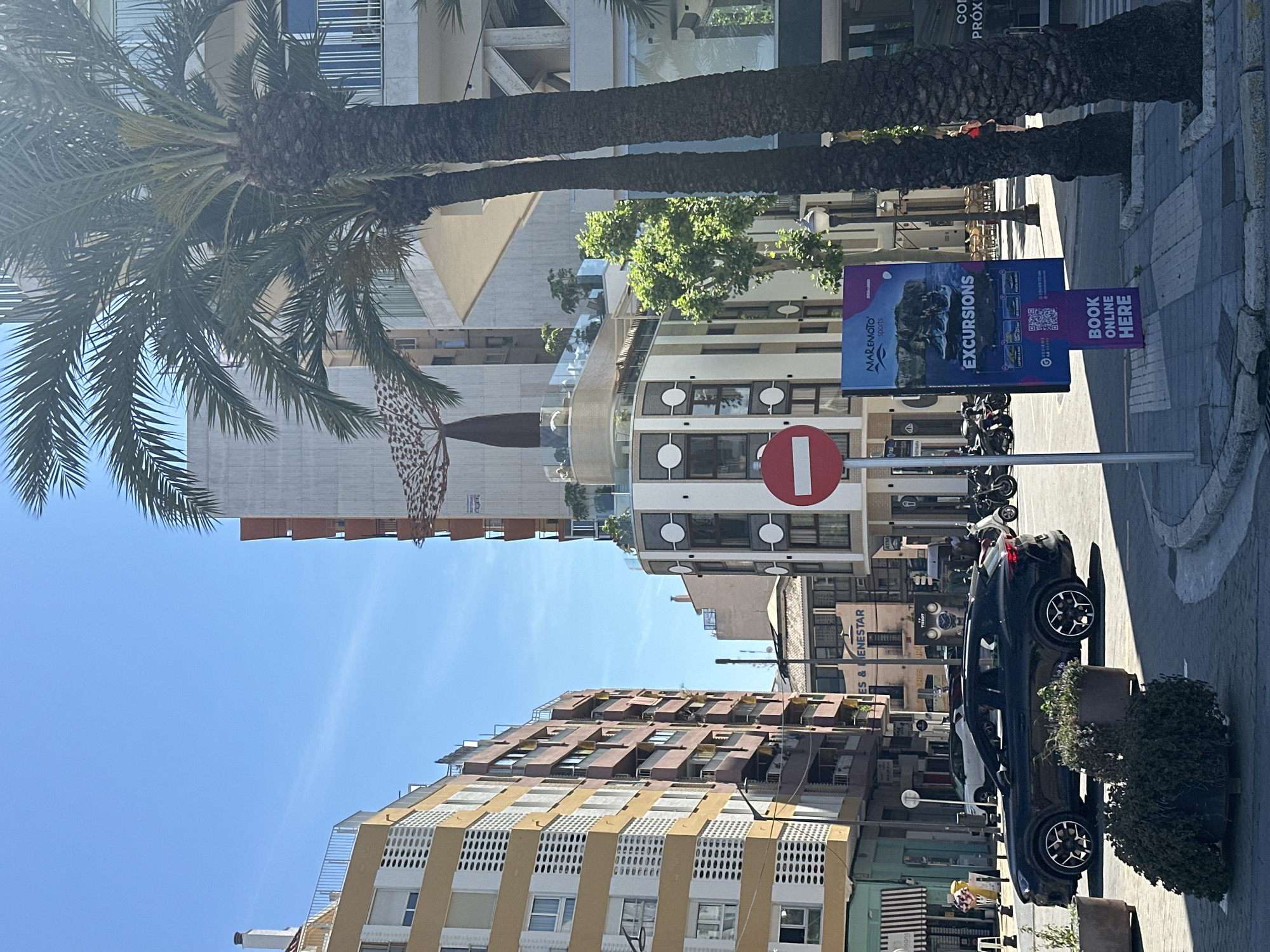 The image shows a street scene in Dénia, Spain. The sky is clear and blue. Buildings of varying heights and architectural styles line the street. The buildings have yellow, white, and brown facades. Palm trees are visible on the left. A "No Entry" sign and an advertising sign for "Maremoto Sports Excursions" are on a post. Several parked motorcycles and a dark-colored car are also seen on the right side of the street. A person in a red shirt is on a building roof. A pot with shrubbery is located beside the vehicle.