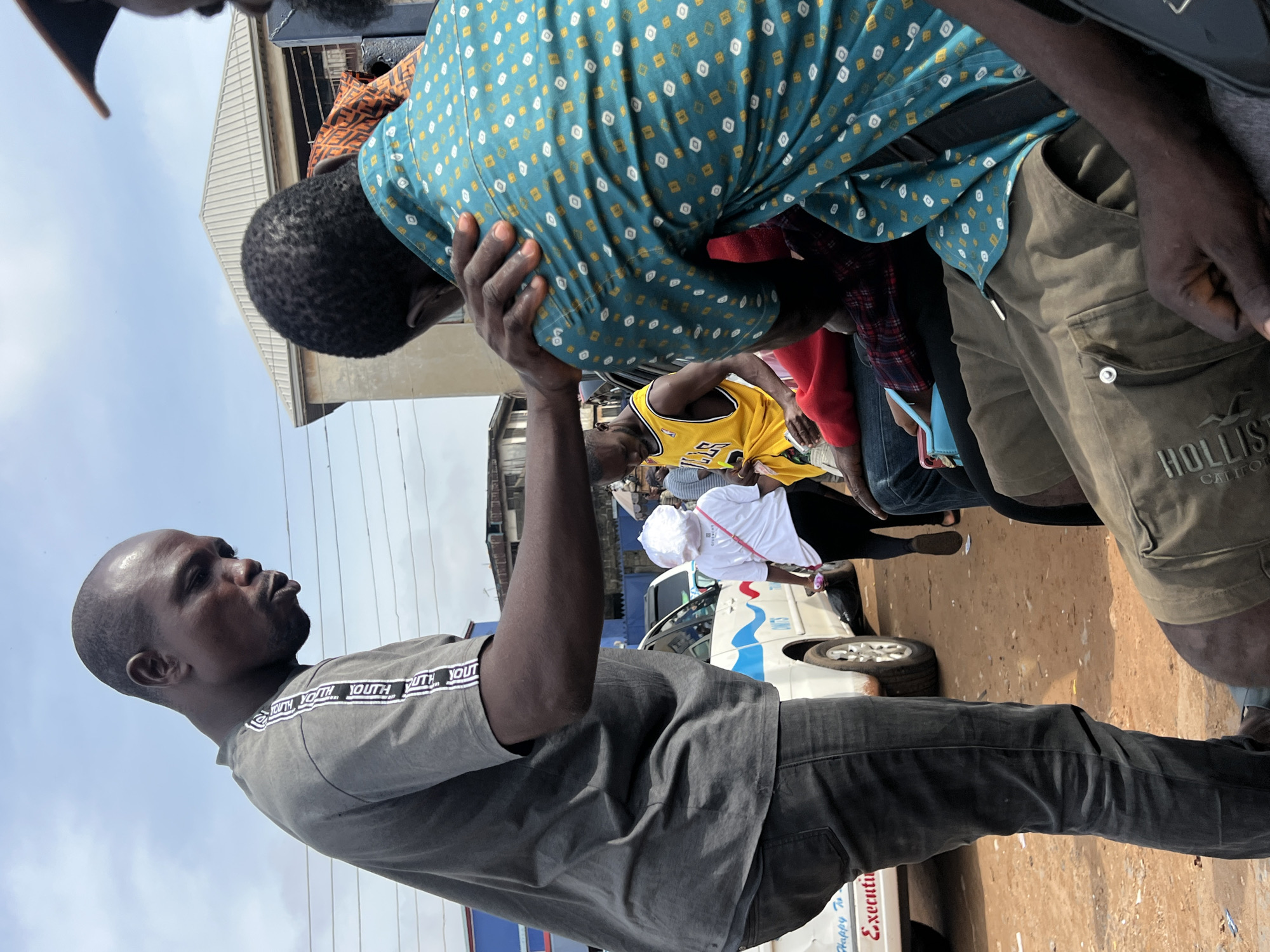 The image captures a scene on a dusty, unpaved street in Onitsha, Nigeria, likely a transportation hub or a busy market area. The overcast sky suggests a humid day. In the foreground, a man wearing a grey t-shirt with white striped lettering on the sleeves is speaking to another man who is seated, wearing a teal shirt with a diamond pattern. The seated man has his hand resting on the shoulder of the man in the grey t-shirt. Behind them, other individuals are visible, some seated and others standing. One person in a yellow basketball jersey is holding a drink and appears to be interacting with others. A white minivan with blue and red stripes is parked nearby, suggesting a commercial transport setting. The overall atmosphere is that of a typical busy day in Onitsha, with the presence of informal transportation and people engaged in everyday activities.