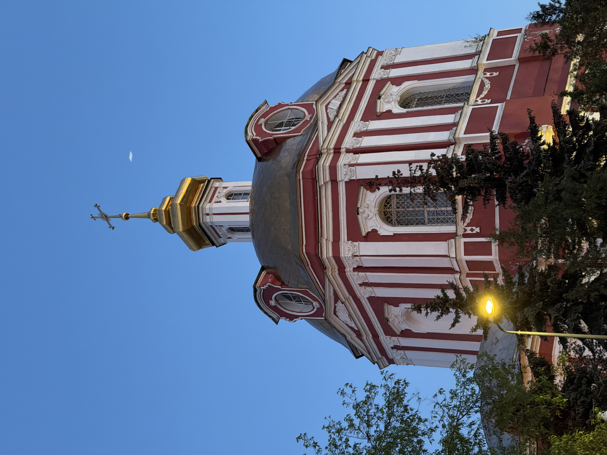 The image shows a portion of the Vysokopetrovsky Monastery in Moscow, Russia. The structure is a red and white building with golden dome. There is a cross on the top of the dome. The building is partially obscured by trees and street light. The sky is blue.