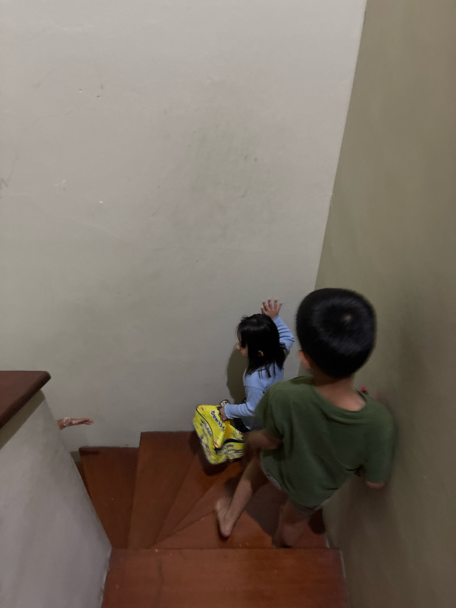 In Bekasi, Indonesia, two young children are seen on a wooden staircase. The child on the right, from behind, wears a green t-shirt and grey shorts, and appears to be leaning against a light brown wall. The child on the left, a girl with dark hair, is positioned further up the stairs. She is wearing a light blue long-sleeved shirt and is holding a yellow bag. Her left arm is raised as if in mid-motion. To the far left of the image, on the wall above the staircase, a child's arm is visible, extended horizontally. The staircase itself is constructed from dark wood with visible treads and risers, leading upwards between two walls. One wall is a light, neutral color, while the other is a darker, earth tone.