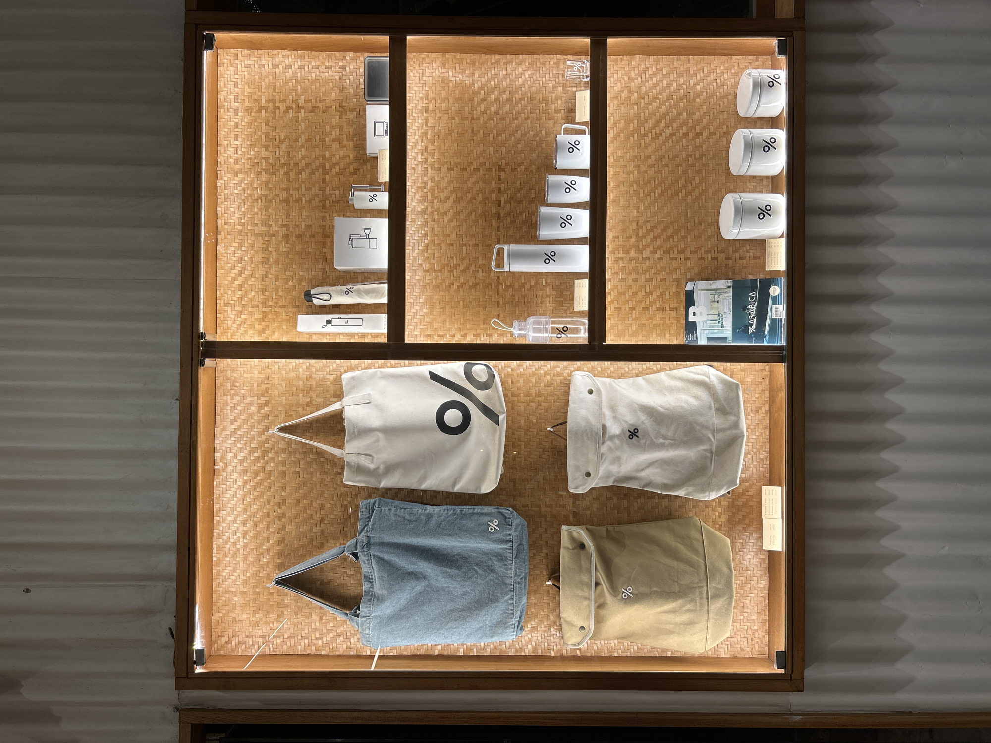 The image shows a retail display window featuring various % Arabica branded merchandise. The display is divided into two vertical sections and has two shelves, all backed by a woven bamboo-like material.
On the left side of the top section, two canvas tote bags are displayed hanging. The left bag is a light blue denim tote, and the right bag is a white tote with a large black "%" logo. Below these, also on the left side, are two more canvas bags, one in a tan color and the other in a cream color, both with a smaller black "%" logo and a flap closure.
The right side of the top section displays several items on a shelf. From left to right, these include two white cylindrical items with black "%" logos (likely water bottles or canisters), a white box with line art and a "%" logo, a small silver item, and a black rectangular object.
The bottom shelf, spanning both sections, also displays merchandise. On the left side, there is a magazine or booklet with "% ARABICA" branding. Next to it are three white cylindrical containers with "%" logos, varying in size. On the far right of the bottom shelf, there are two white cups with "%" logos and a small clear glass with a "%" logo.
The display is lit from above by LED strip lights, casting a warm glow on the items. The overall aesthetic is minimalist and modern. The retail space appears to be located in Kuta Selatan, Indonesia, with a corrugated metal wall visible in the background.