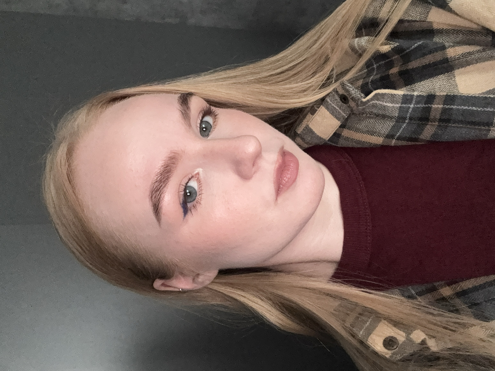 A young woman with blonde hair is posing for a selfie or photo. She is wearing a maroon sweater underneath a tan and gray plaid flannel shirt. She has fair skin and noticeable makeup, including blue and white eyeliner on her lower eyelids. She has an earring in her left ear. The background is a dark gray or black color. There is no indication of the location being Chaikovsky, Russia or any specific city.