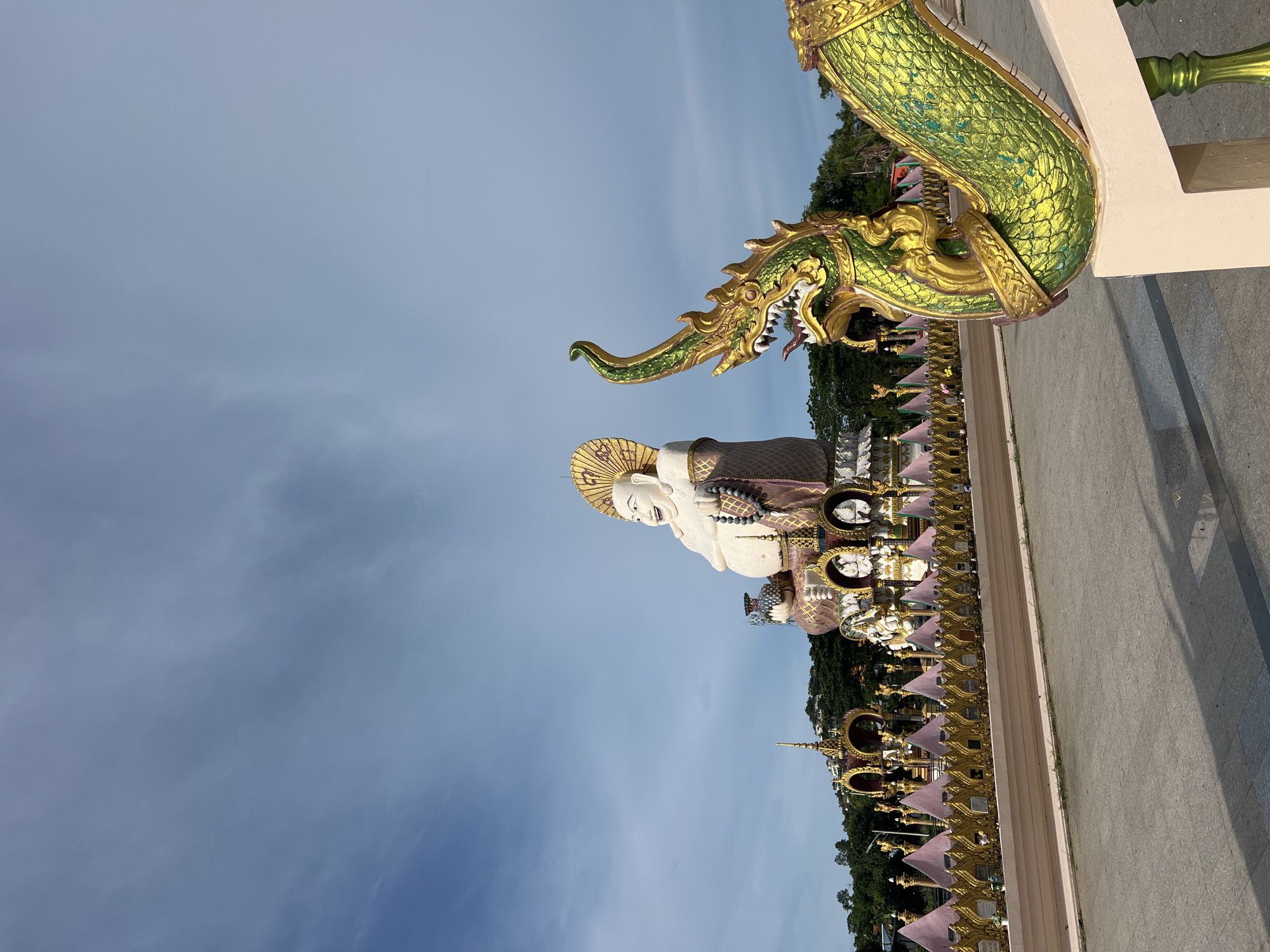 The image shows a large statue of a smiling Buddha in the background, at Baan Plai Laem Temple in Koh Samui, Thailand. In the foreground, a large, ornate green and gold serpent statue is visible, its head and part of its body curving towards the right of the frame. The serpent statue appears to be a decorative railing or support. Behind the serpent statue and in front of the Buddha, there is a golden fence with pink triangular accents. The ground is paved, and a shadow is cast from the serpent statue. The sky is blue with scattered clouds. The overall scene depicts a religious and cultural site with elaborate statues and decorations.
