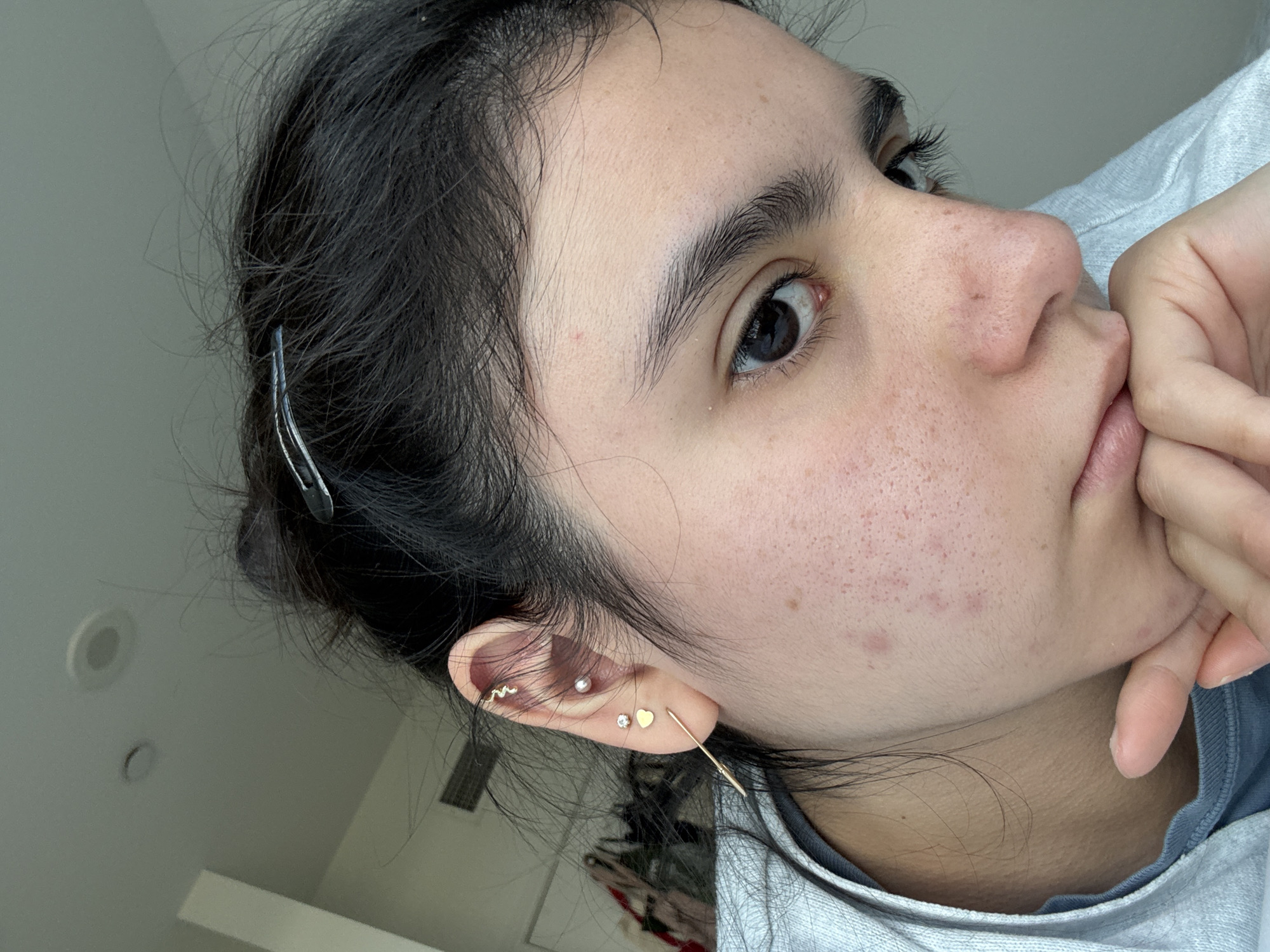 A close-up, slightly tilted portrait captures a young person, likely female, with dark hair pulled back in a casual style, possibly in Austin, United States of America. Loose dark hair strands frame the face. The subject's dark eyes gaze directly towards the viewer. Their skin shows natural texture with scattered freckles or hyperpigmentation on the cheeks and nose. The chin rests gently on the back of their right hand, with fingers lightly touching the lips, suggesting a pensive or relaxed posture.
The person wears a light gray casual top, possibly a sweatshirt. The visible left ear features multiple gold-toned piercings: a small heart stud, a clear crystal stud, a small pearl stud, and a curved bar or snake-like earring in the helix area. A longer, thin gold earring dangles from the earlobe. A simple dark hair clip is visible in the pulled-back hair on the left side of the head. The background is a soft-focus, light-toned indoor setting, with glimpses of a white ceiling and two circular recessed lights. The lighting is soft and even, indicating daytime. There is no visible text.