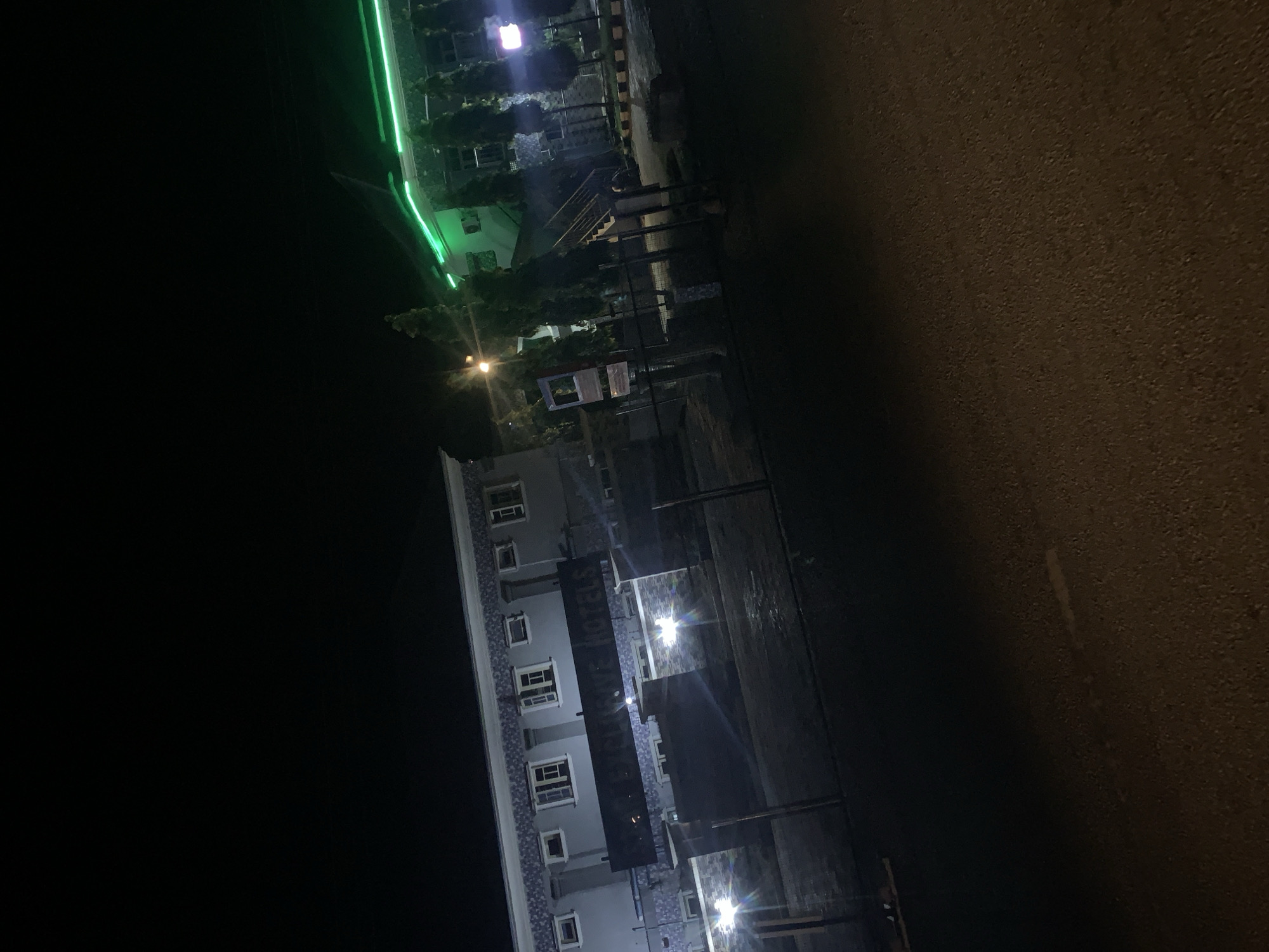 This is a nighttime outdoor photograph of a hotel in Jalingo, Nigeria. The hotel appears to be a multi-story building with a dark facade and multiple windows. A large sign is mounted on the front of the building, but the text is partially obscured. The hotel is illuminated by exterior lighting. To the right, another section of the hotel is visible with a green neon light strip running along its roofline. Several trees are planted in front of this section. The ground in front of the hotel is paved, and a fence runs along the perimeter. The sky is dark, indicating it is nighttime.