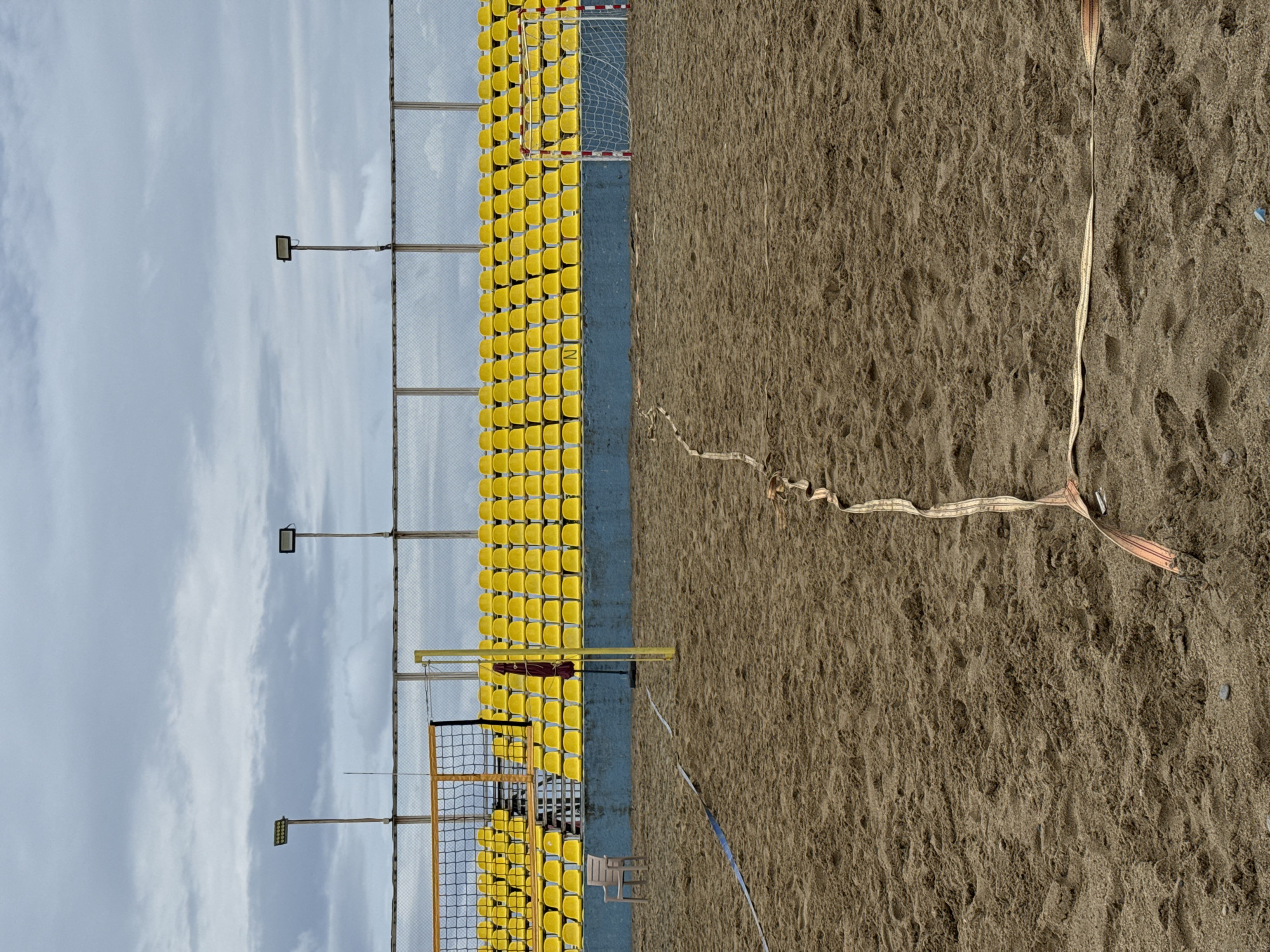 The image depicts an empty outdoor beach volleyball court in Batumi, Georgia, under an overcast sky. The sandy court features boundary lines marked with beige and blue straps, with a volleyball net spanning the center. In the background, a tiered stadium seating area with bright yellow seats is visible, backed by a wire mesh fence and topped with a railing. Two tall floodlights are positioned above the stands. A single plastic chair sits near the net on the left side of the court, and a red umbrella hangs on the yellow net post. A small goal net, typically used for beach soccer or handball, is present on the right side of the court, behind the yellow seating. The scene is devoid of people.