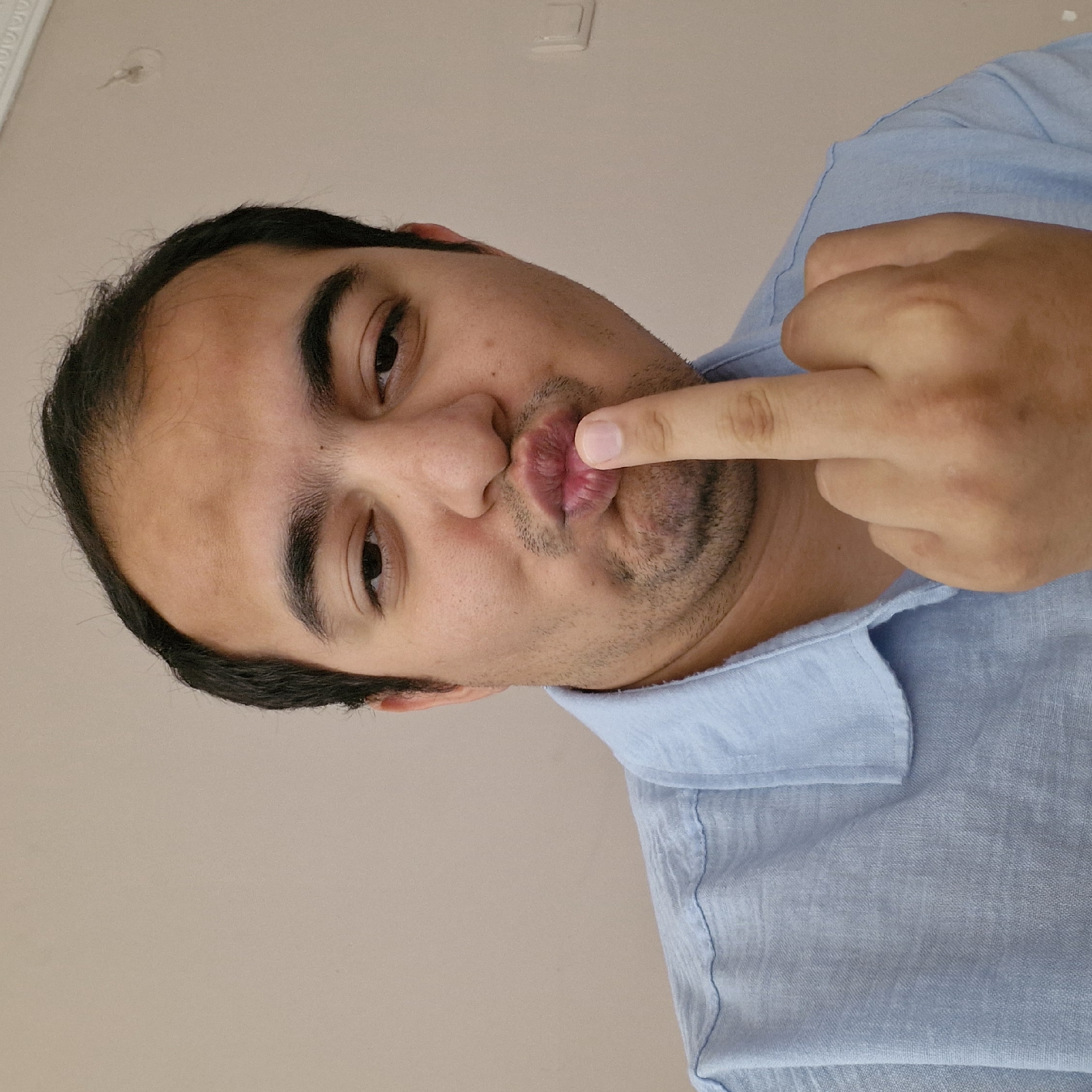 A close-up, waist-up shot shows a man with dark hair and eyebrows, a light complexion, and a thin mustache. He is wearing a light blue collared shirt. His lips are pursed as if he is about to kiss or give a blow kiss. His right index finger is touching his lips. He is facing the camera and is positioned in front of a plain, light-colored wall. The scene is indoors, and no specific location in Küçükçekmece, Turkey can be determined from the image.