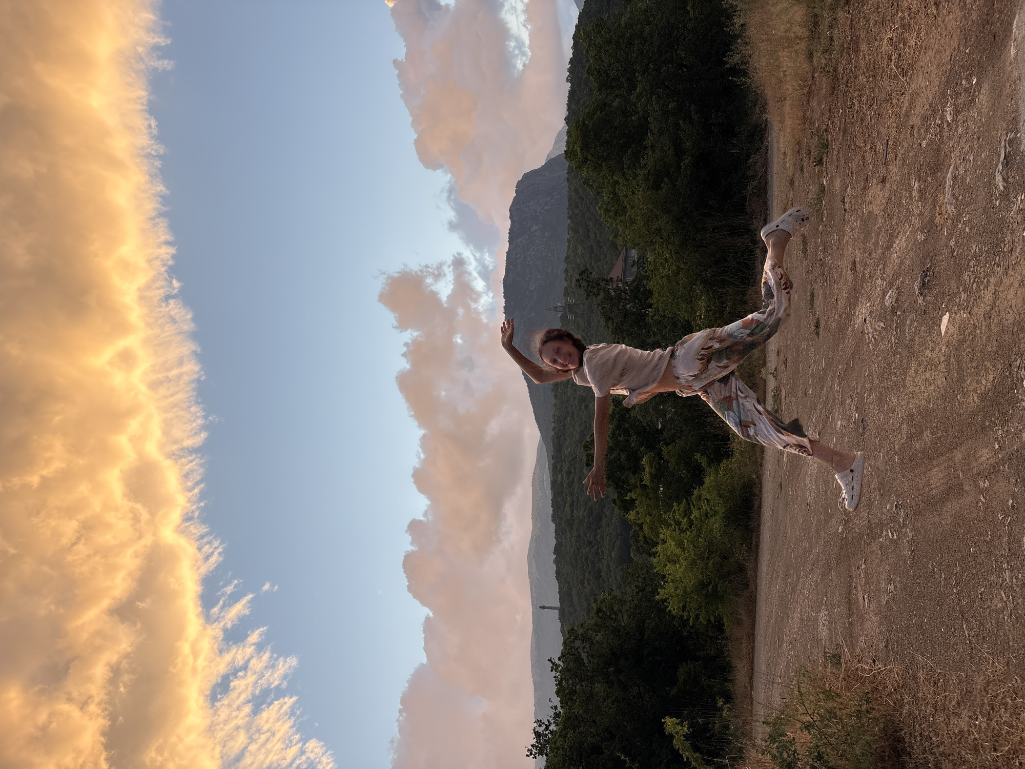 A young woman is captured mid-jump on a dirt road with a scenic mountain and cloud-filled sky backdrop. The woman is wearing a light-colored shirt and patterned pants, with her arms outstretched in a dynamic pose. The surrounding environment includes lush green trees and rolling hills, with a tower visible in the distance on one of the mountains. The sky is illuminated by the warm hues of sunset, casting a golden glow on the clouds. The location is the city of Malyi Mayak, Ukraine.