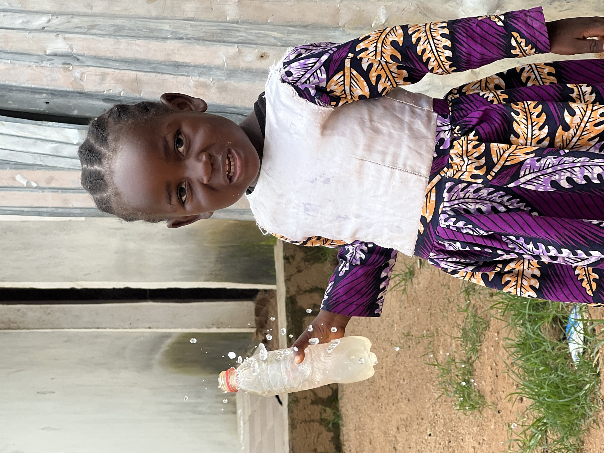 A young African girl is standing outdoors in what appears to be a residential area in Jos, Nigeria. She is wearing a white top with a colorful patterned skirt and sleeves featuring purple, orange, and black leaf designs. In her left hand, she holds a clear plastic bottle filled with a light-colored liquid, from which water is splashing and dripping. The background consists of a textured wall and a corrugated metal fence. The ground is covered with dirt and patches of grass.
