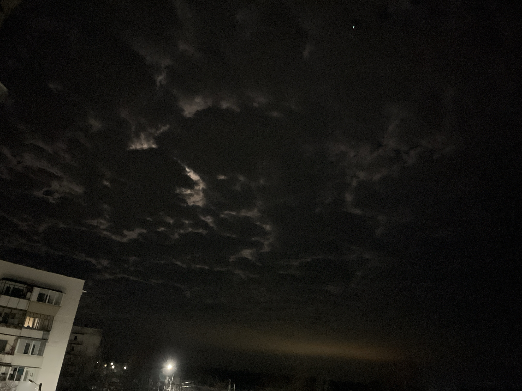 The image shows a nighttime scene in what appears to be the city of Chaikovsky, Russia, based on the architecture. The sky is dominated by dark, heavy clouds with some patches illuminated by the moon. A multi-story residential building is visible on the left side of the frame, with some windows lit. In the distance, a street lamp illuminates the area, casting a glow on the horizon. The overall environment is dark and suggestive of an urban setting.