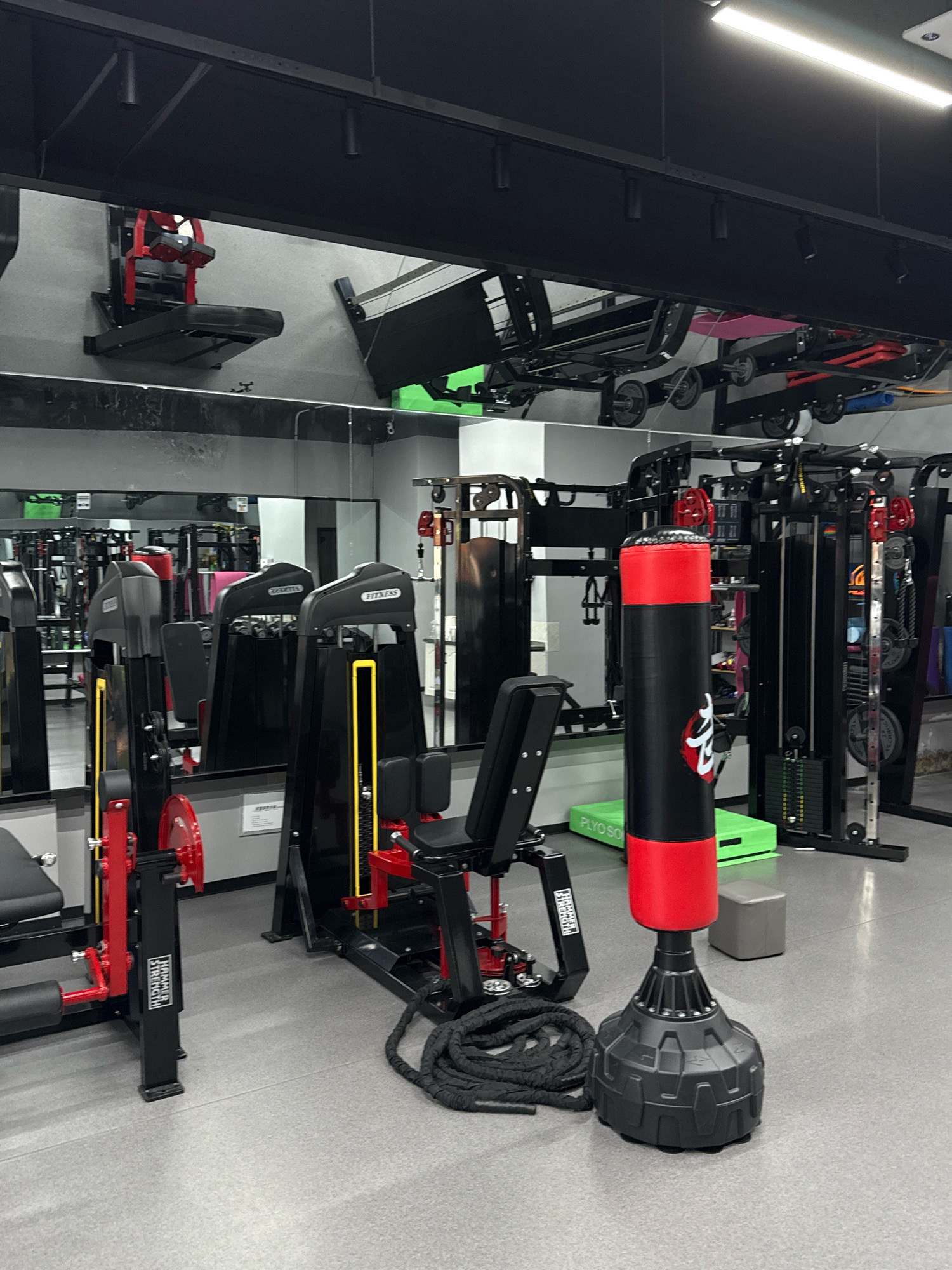 The image shows the interior of a gym, likely located in Yiwu, China, given the context. The gym features a variety of fitness equipment, including weight machines, a punching bag, and what appears to be a battle rope coiled on the floor. The equipment is predominantly black and red, with some yellow accents on the weight machines. The space is well-lit with fluorescent lighting from the ceiling. Mirrors are strategically placed on the walls, reflecting the gym equipment and creating a sense of a larger space. The floor is a light gray, speckled material. No people are visible in the image, suggesting the gym is either empty or the photograph was taken during off-hours. The overall impression is one of a modern, functional fitness facility.