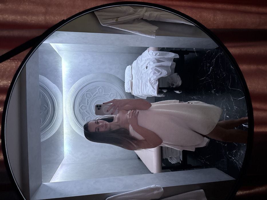 A young adult female is depicted in a mirror selfie, wrapped in a white towel that covers her from just above her breasts to mid-thigh. She holds a light-colored smartphone with her right hand, obscuring part of her chest, while her left hand rests on the towel near her chest. She has long brown hair, dark manicured nails, and a slight smile.
The reflection reveals an interior room resembling a spa or massage therapy treatment area. The room features light grey walls and ceiling, illuminated by indirect strip lighting, and a dark, veined marble floor. Two prominent circular white bas-relief panels with intricate, mandala-like patterns are affixed to the far wall. Two massage tables are visible: one on the left, partially obscured, with a stack of neatly folded white towels beneath it, and another on the right, covered in white, somewhat rumpled linens. A white bathrobe hangs on a hook or rack to the far right within the reflection. A portion of another white towel is visible hanging on a hook to the left of the reflected room. The mirror has a black frame, and a reddish-orange curtain is partially visible outside the mirror's frame on the right.
The exact location within the city of Kyiv, Ukraine, cannot be determined from the provided image.