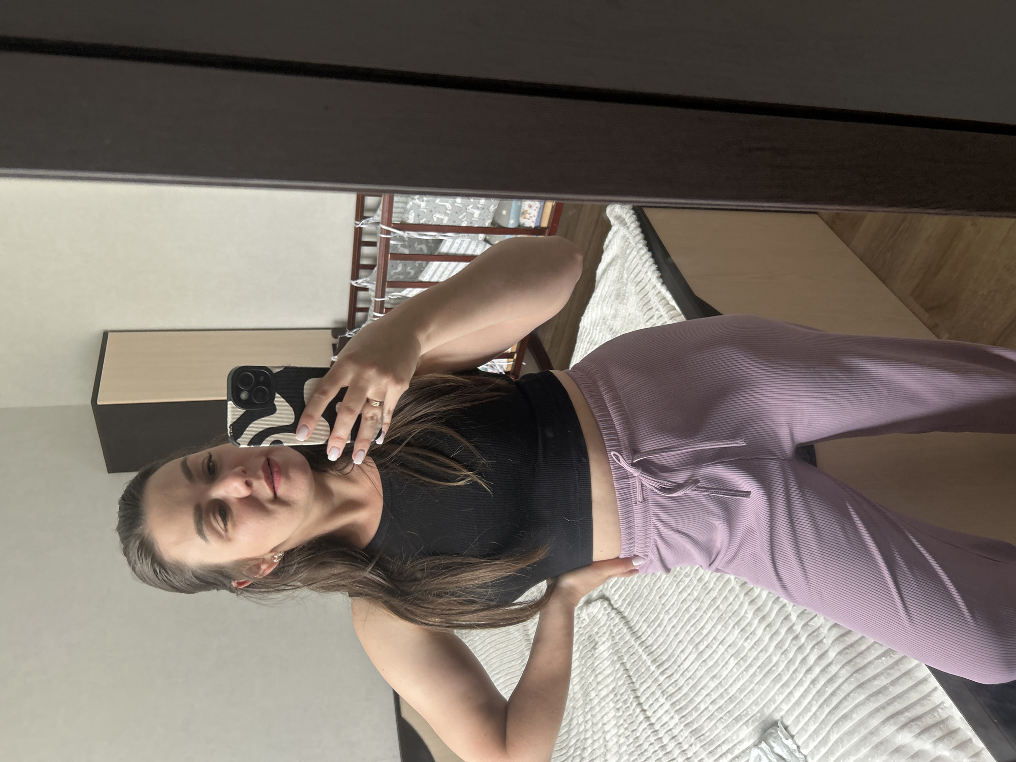 A woman is taking a selfie in a mirror inside a room. She is wearing a black ribbed crop top and light purple ribbed pants with a drawstring. The room appears to be a bedroom with a white comforter on the bed. A crib with decorative elements and a dresser or cabinet are visible in the background, reflected in the mirror. The location could be a residence in Chaikovsky, Russia.