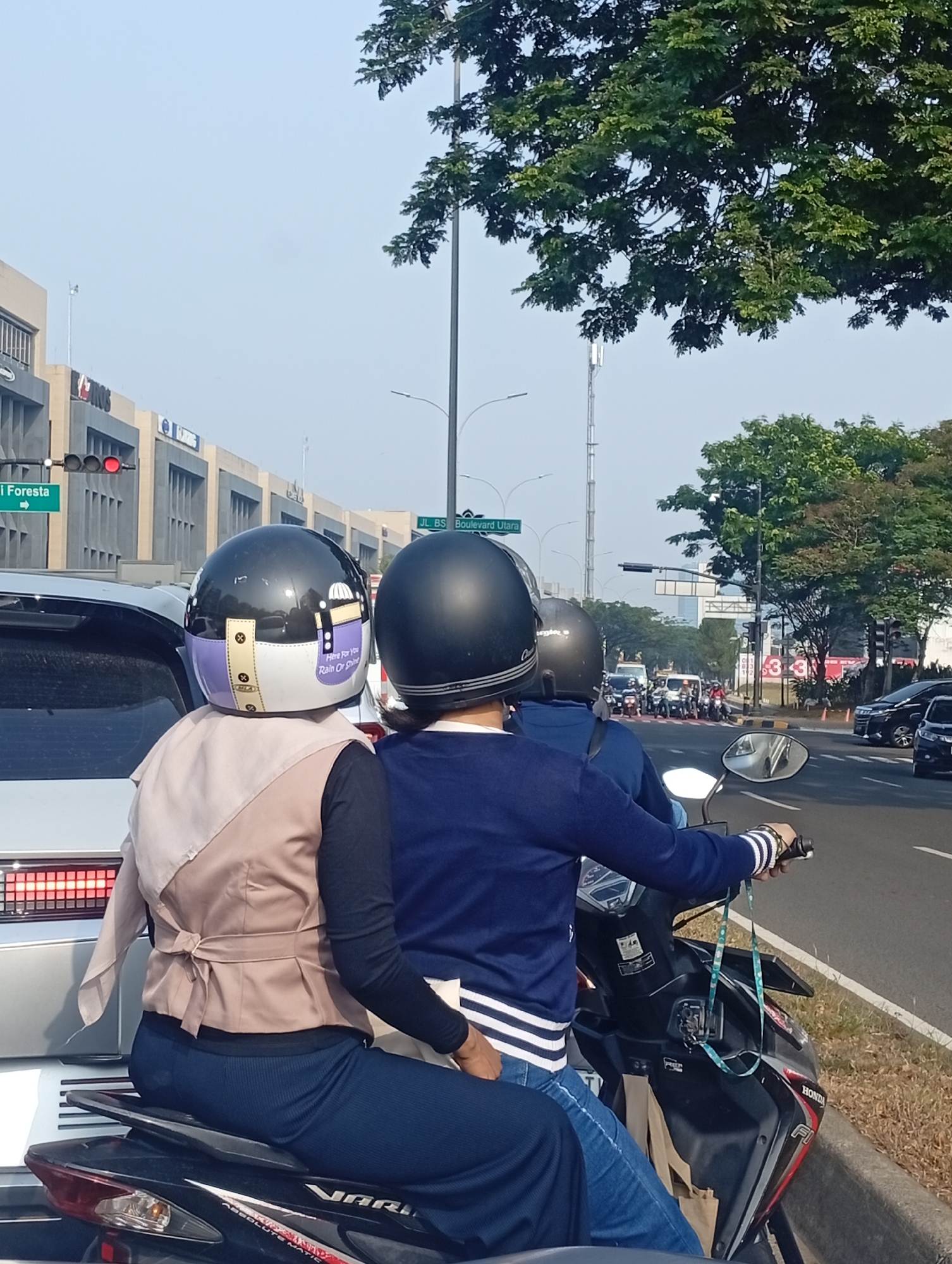 Two individuals are seated on a scooter in traffic on a street in Lengkong Kulon, Indonesia. The person in the front wears a black helmet with purple and white accents and a beige vest over a black long-sleeved shirt. The person behind wears a black helmet and a dark blue long-sleeved shirt with white stripes on the cuffs. Both are looking forward. The scooter has a black seat and a black and red body. In the background, there are buildings, trees, a traffic light displaying red, street signs, and other vehicles including cars and motorcycles. The sky is clear and blue. The scene appears to be a street with commercial buildings and moderate traffic.