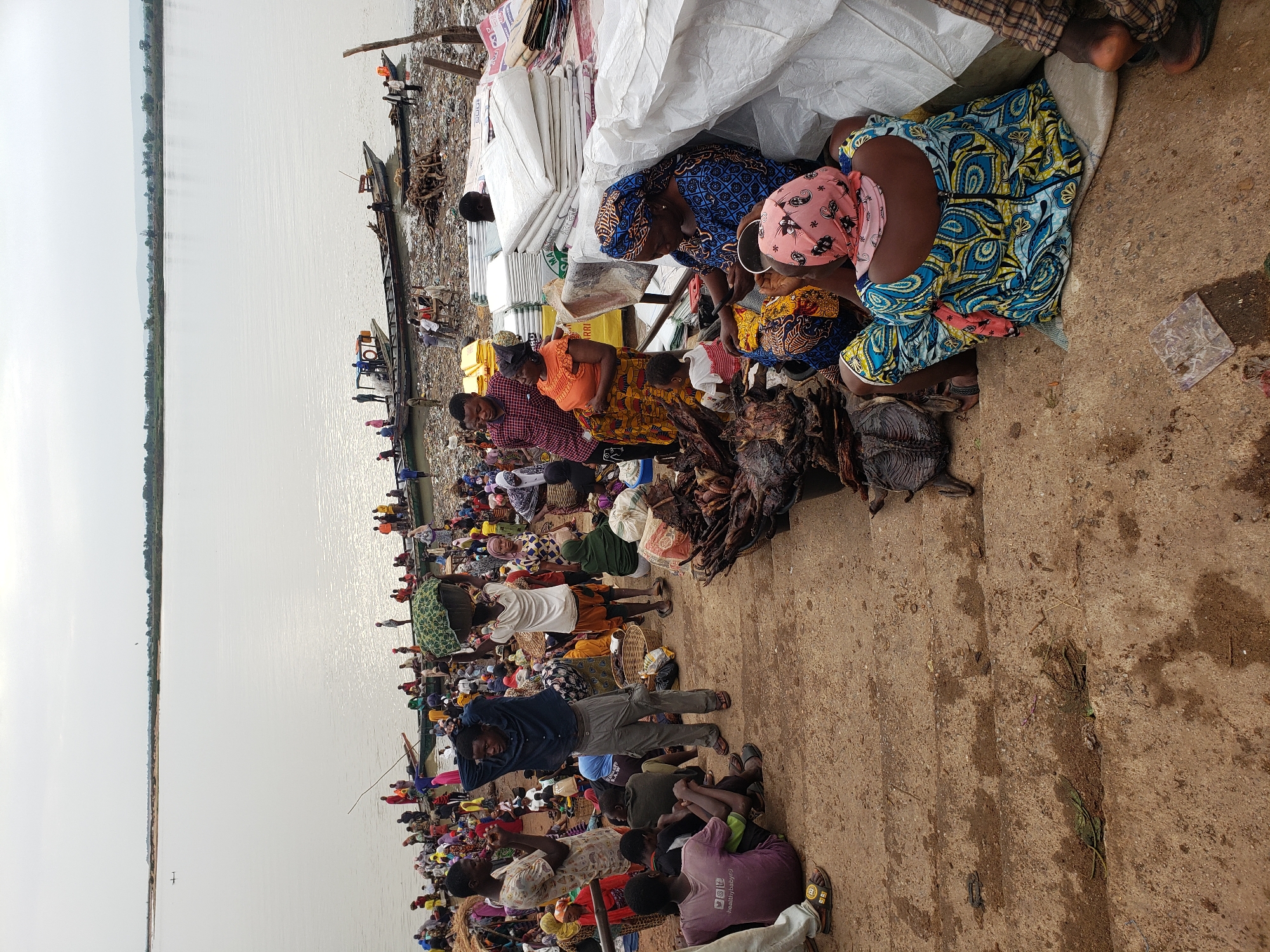 This image depicts a bustling riverside market scene in Lokoja, Nigeria. People are densely packed along the shore, with many sitting on the ground amidst piles of goods. Several boats are docked along the river's edge. White sacks of goods, possibly produce, are visible. Many people wear colorful clothing. Some individuals are sitting and appear to be selling or preparing items. There are piles of what appears to be dried fish or meat. The ground is bare earth. The sky is overcast, indicating a potentially humid environment.