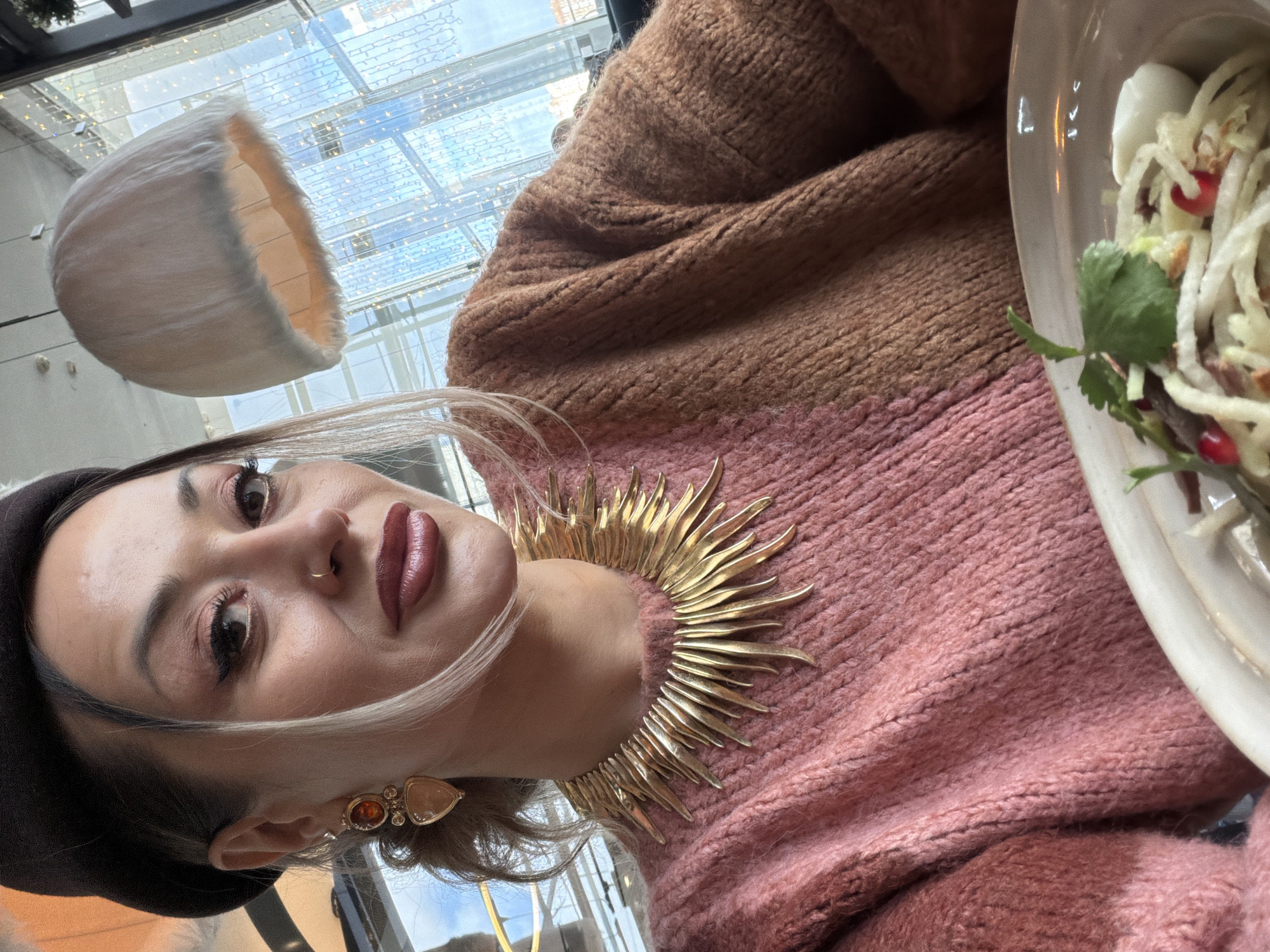 A woman wearing a black beret, a two-tone brown and pink sweater, and a gold, sun-shaped necklace is seated at a table. Her left ear has a decorative, multi-stone earring. To her right is a plate of food that appears to contain shredded white vegetables or noodles, green herbs, and pomegranate seeds. In the background, a partially visible, white, lamp-like object hangs near a glass ceiling with lights that suggest the city is Krasnoyarsk, Russia.