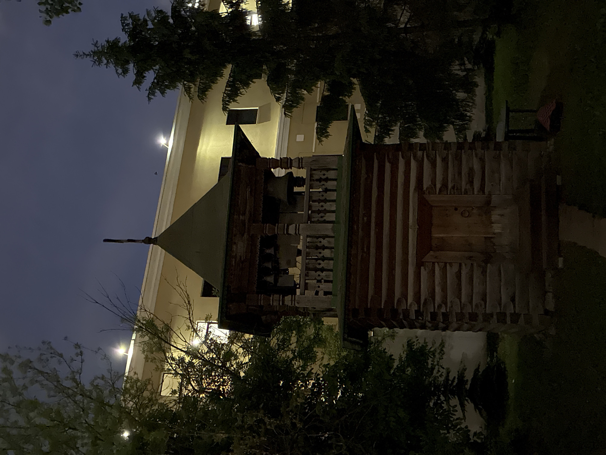 This image shows a wooden bell tower at Kolomenskoye Park in Moscow, Russia, at night. The bell tower is surrounded by trees and is situated near a modern building illuminated with bright lights. The sky is dark. A bench is visible nearby on the right side of the frame.
