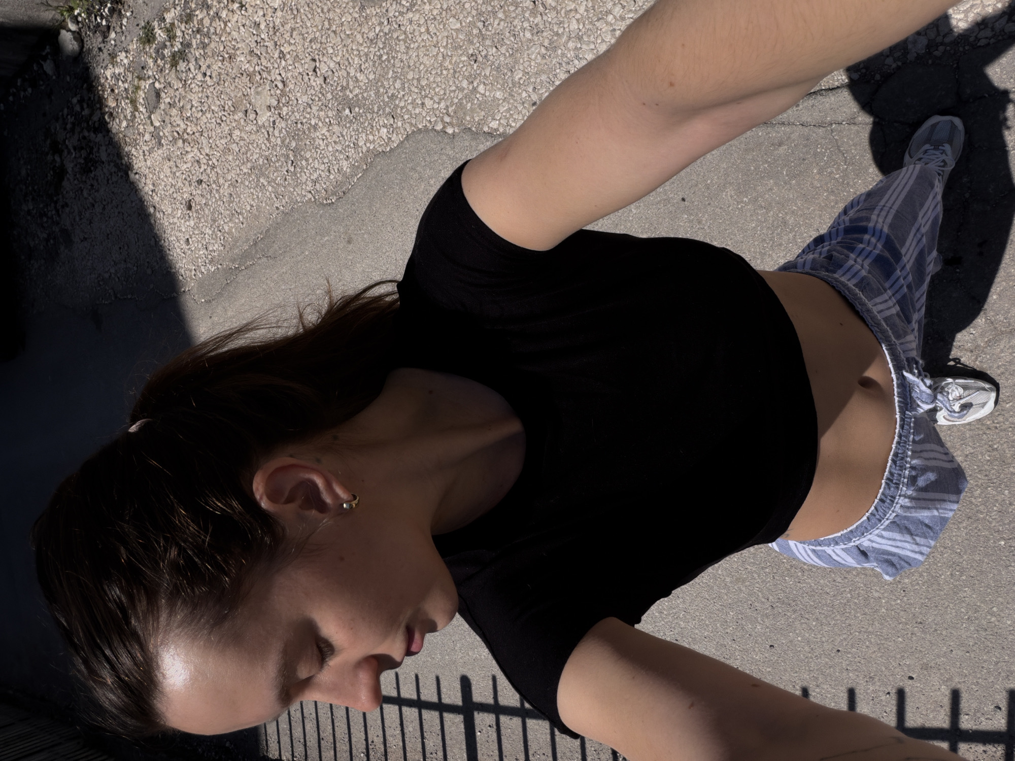 A high-angle outdoor shot captures a person, likely female, from an overhead perspective. The individual has dark hair pulled back and is looking downwards with their eyes closed or cast low. They are wearing a black short-sleeved top that exposes their midriff, paired with light blue and white striped or plaid shorts and grey athletic shoes. A small gold hoop earring is visible in their right ear. Their arms are extended upwards, suggesting they might be holding the camera for a selfie or being photographed from above.
The setting appears to be an outdoor paved area, possibly a path or sidewalk, with a mixture of concrete and light-colored gravel visible on the ground. Strong shadows are cast on the ground from the person and a dark fence or railing, indicating bright sunlight, likely during mid-morning or afternoon. The overall impression is casual and relaxed in a sunny environment in Budva, Montenegro. No visible text is present in the image.