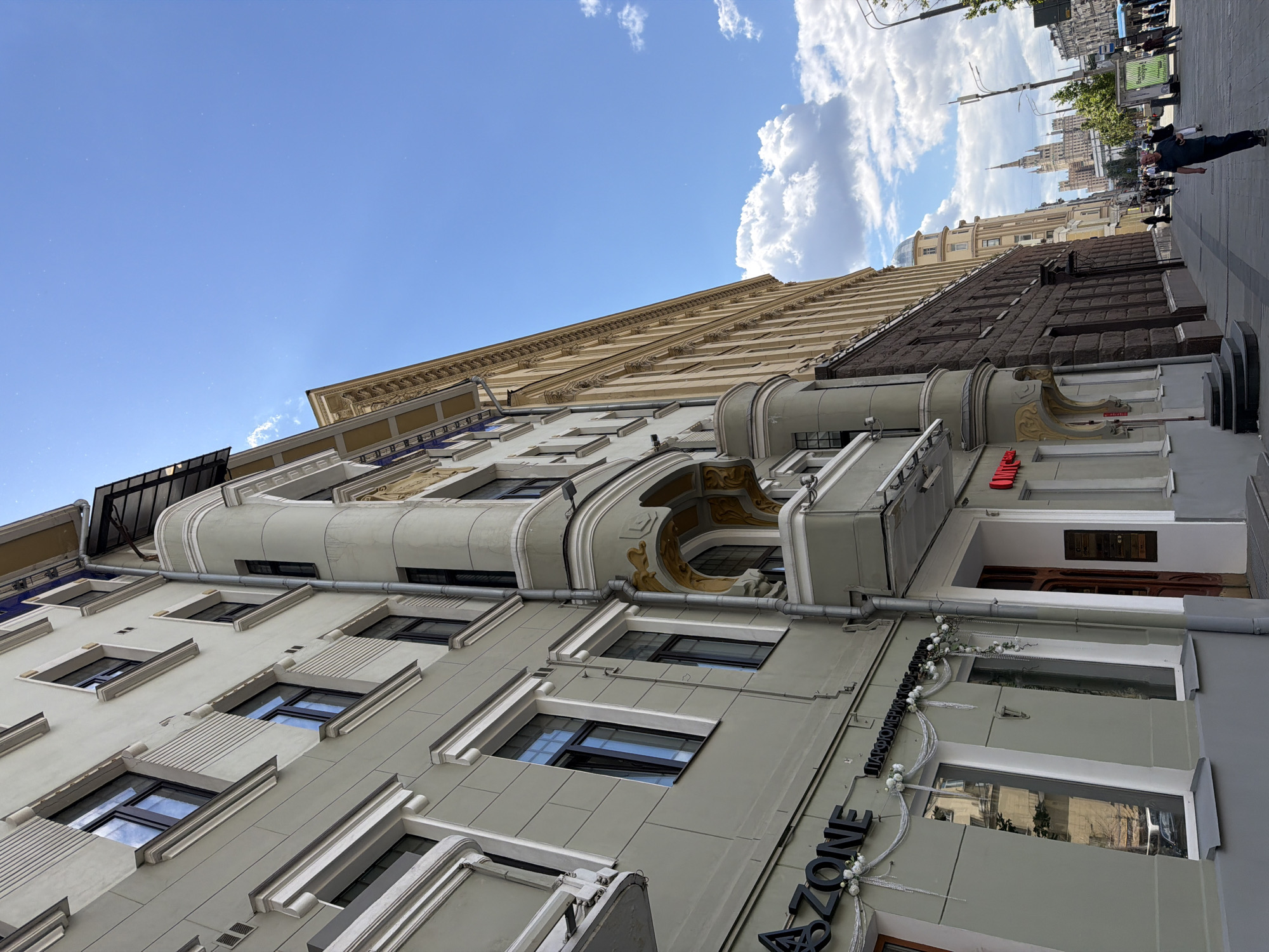 The image presents an upward-angled view of an urban street scene featuring a row of buildings under a bright, partly cloudy sky. The perspective is rotated clockwise, making the buildings appear to lean and the street to ascend towards the right.
In the foreground, a multi-story building with a light greenish-grey facade dominates the left side. It exhibits classical architectural elements, including intricate moldings, cornices, and decorative panels around its numerous rectangular windows. Some sections feature rounded bay windows or small balconies. On the lower right of this building, a black sign reads "ZONE," accompanied by Cyrillic script, likely "ПАРФЮМЕРИЯ" (Perfumery), and adorned with white floral garlands. Above it, a red sign shows part of the word "RED."
Behind the foreground building, another structure with a darker, rough-textured facade is visible. Further down the street, various buildings, including one with a dome, lead to a distant prominent skyscraper characterized by its distinctive spire, indicative of Stalinist-era architecture.
The sky is a clear blue with scattered white, puffy clouds, suggesting a sunny daytime scene. A paved sidewalk runs along the right side of the frame, where several pedestrians are visible. One man in a dark shirt and pants is prominently seen in the mid-ground. Streetlights and other urban infrastructure are also present along the street, indicating an active city environment.