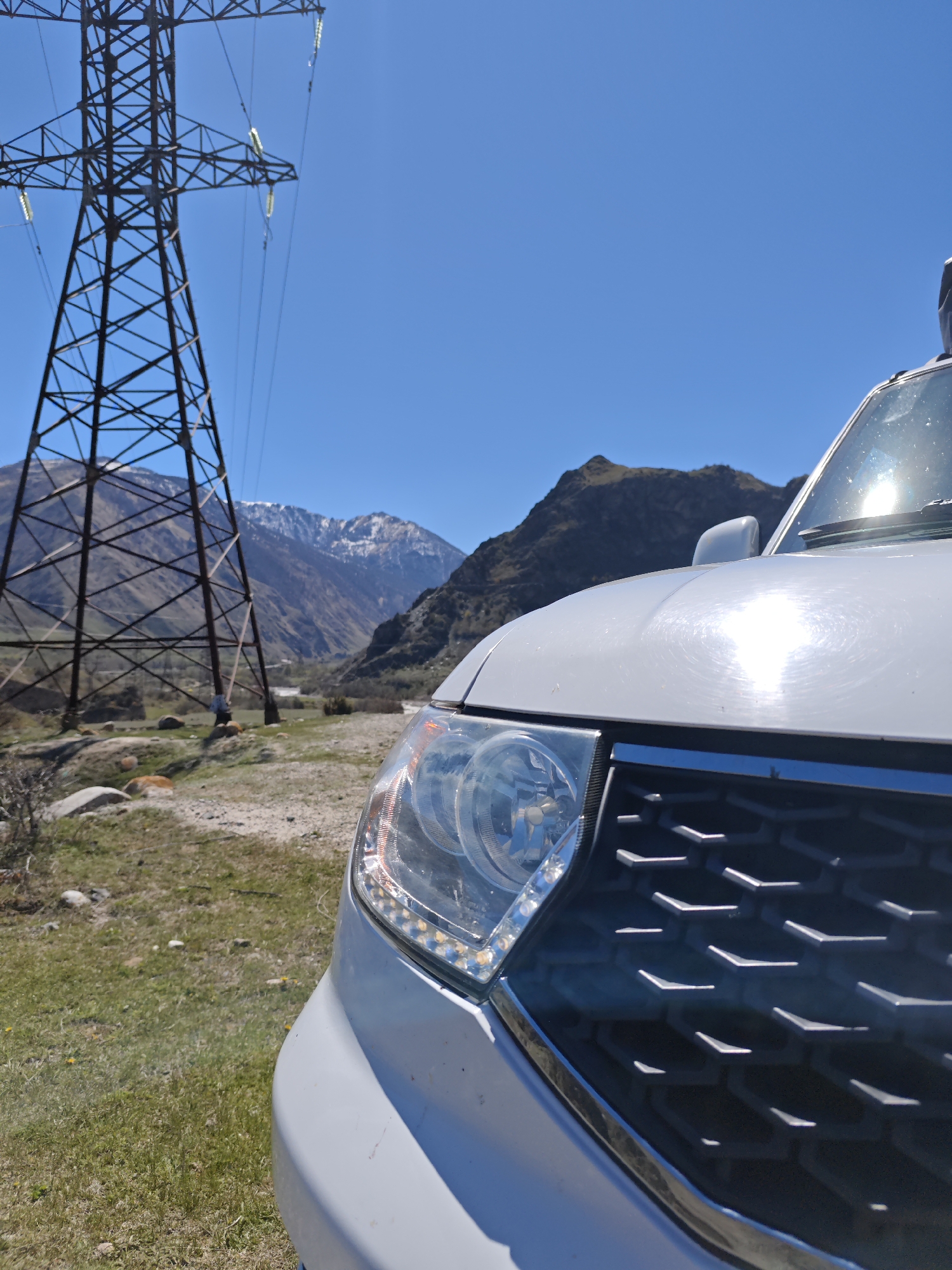 A close-up, outdoor shot features the front right corner of a white vehicle against a mountainous backdrop in Bylym, Russia. Dominating the left side of the frame is a large, metal electricity pylon. Behind the pylon, a mountain range is visible, some peaks capped with snow. A valley stretches between the mountains, appearing green. The sky is a clear blue. The vehicle's headlamp is prominently displayed, along with a portion of the grill. The ground is grassy and rocky. A man stands near the pylon. The sun is shining brightly, reflecting off the vehicle's hood.