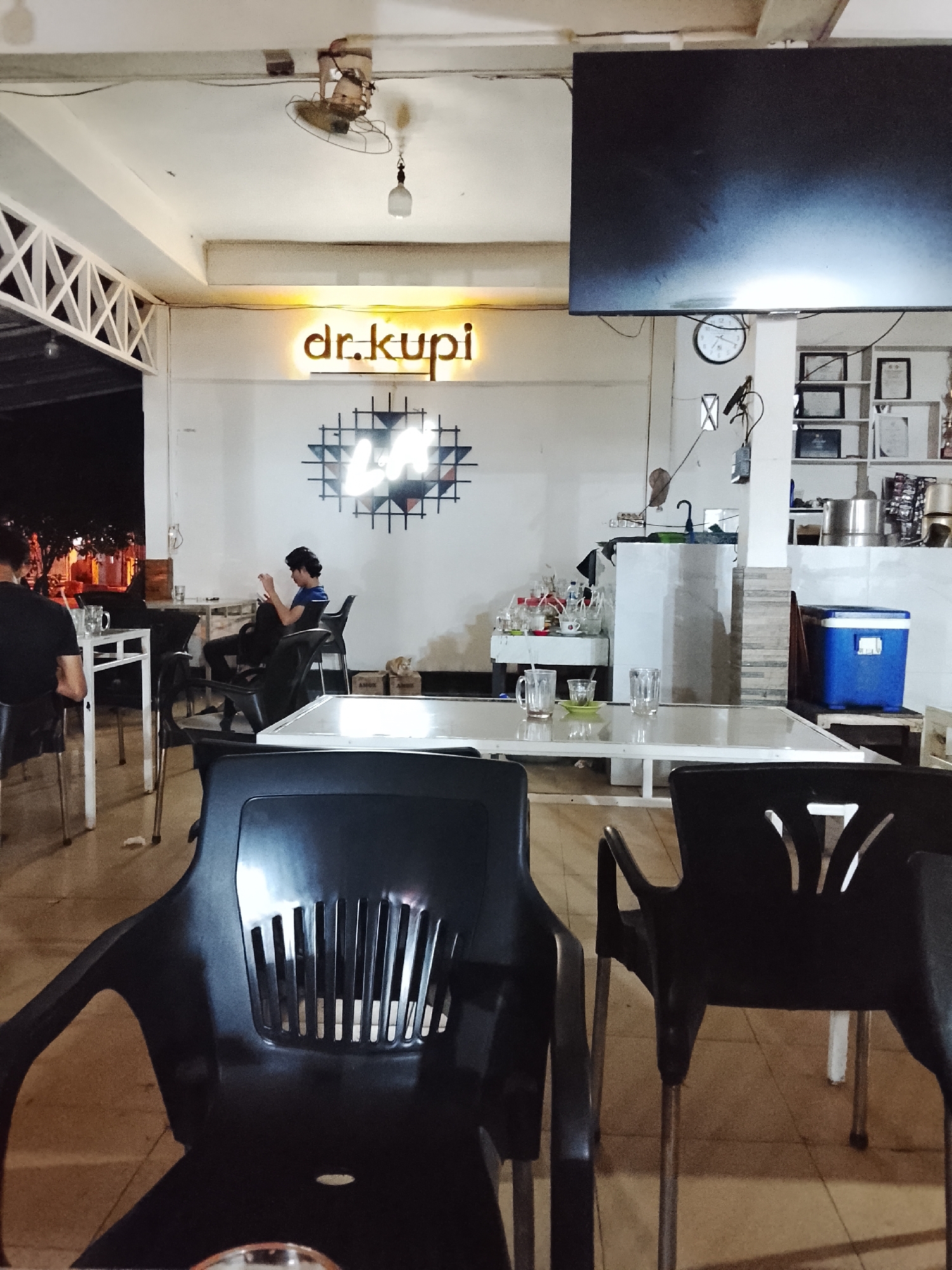 This image captures the interior of a coffee shop named "dr.kupi" in Lhokseumawe, Indonesia, at night. The lighting is dim, creating a cozy ambiance. In the foreground, two black plastic chairs are visible, one in focus and the other slightly blurred. Further back, a man is seated at a table, looking at his phone. The coffee shop features white walls adorned with neon signs, including the "dr.kupi" logo and an abstract geometric design. Several tables and chairs are arranged throughout the space, and a television screen is mounted on the wall. A blue cooler box sits in the background, near shelves stocked with various items. The overall scene depicts a typical evening at a relaxed coffee shop.