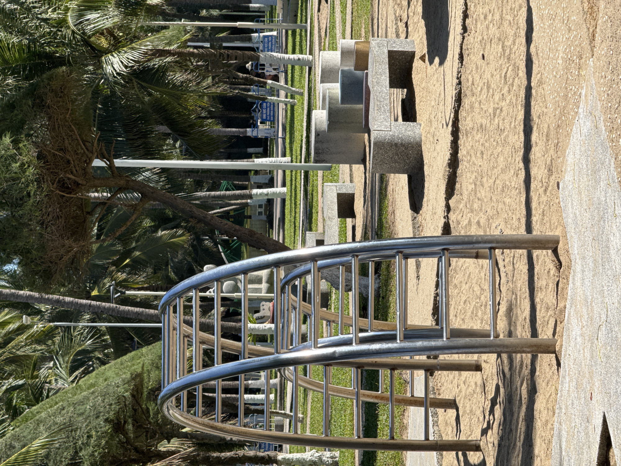 The image depicts an outdoor recreational area within a park or promenade in Nha Trang, Vietnam, under clear, sunny conditions. In the foreground, a prominent stainless steel arched monkey bar structure stands on a sandy surface, with multiple horizontal rungs. To the right, a cluster of modern-style concrete or granite seating elements is arranged on the sand, consisting of various cylindrical stools and rectangular benches, some featuring colored tops. The sandy area gives way to a concrete perimeter in the immediate foreground.
The midground features well-maintained green grass interspersed with numerous trees, including tall palm trees and other tropical species, many with white-painted lower trunks. Multiple white light poles are visible throughout the park. In the background, behind a row of trees, blue barriers with white Vietnamese text, including "KHU VỰC CẤM" (Restricted Area), mark a boundary. A single individual, obscured by distance and wearing a light-colored wide-brimmed hat, is visible walking towards the right, behind these barriers. The scene is brightly lit, casting distinct shadows from the structures and trees onto the sandy and grassy grounds. No direct interactions or specific activities are observable beyond the general setting.