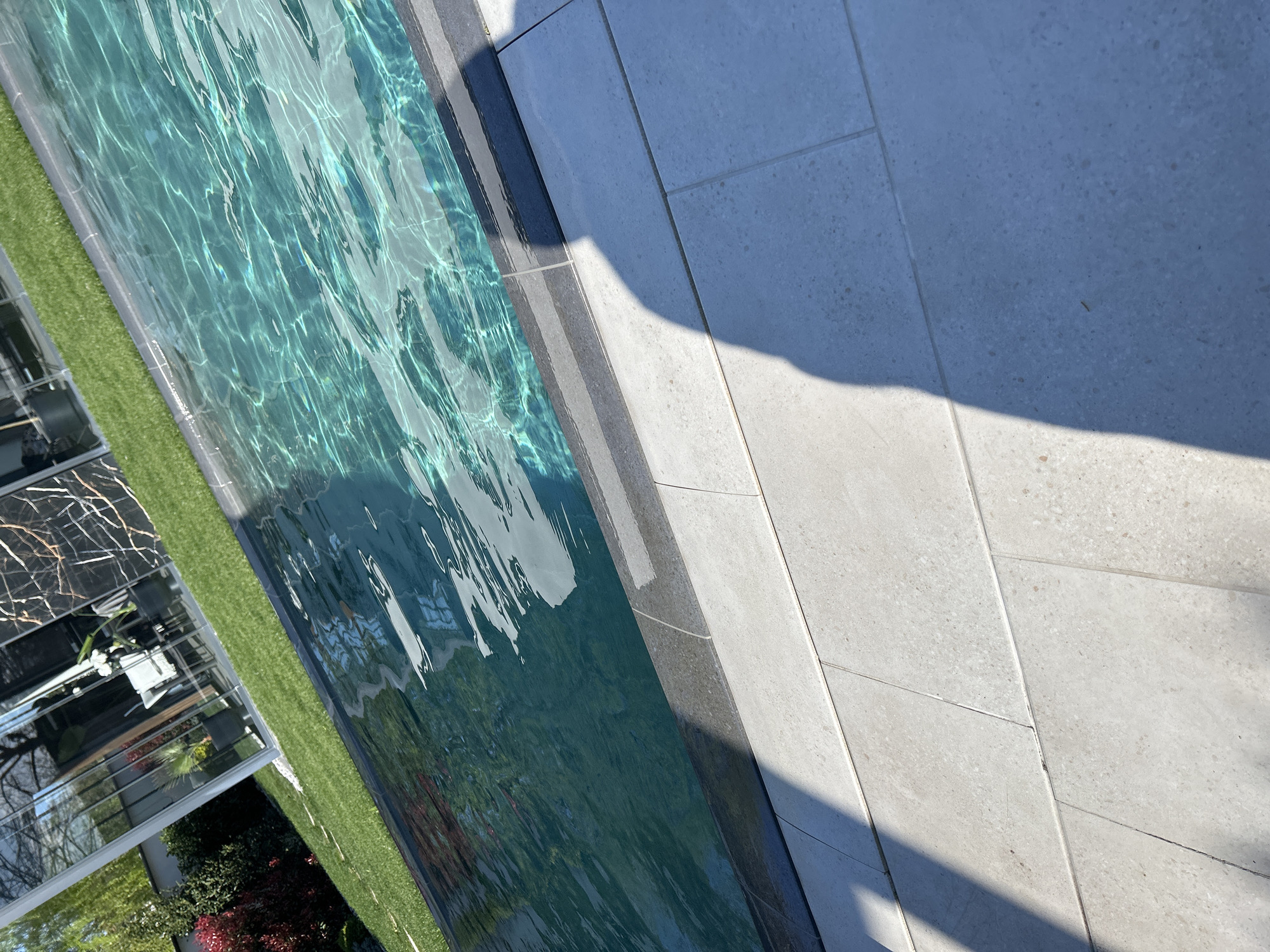 The image depicts a section of a swimming pool adjacent to a tiled surface, possibly a patio, with an artificial turf area and parts of a building visible in the background. The pool water is clear and reflects the surrounding environment. The tiles are light gray and appear to be made of stone. A shadow falls across the tiles. Part of a building with glass windows and possibly a balcony is visible beyond the turf, along with some greenery. The location is stated as Plovdiv, Bulgaria, but no details in the image explicitly confirm this.