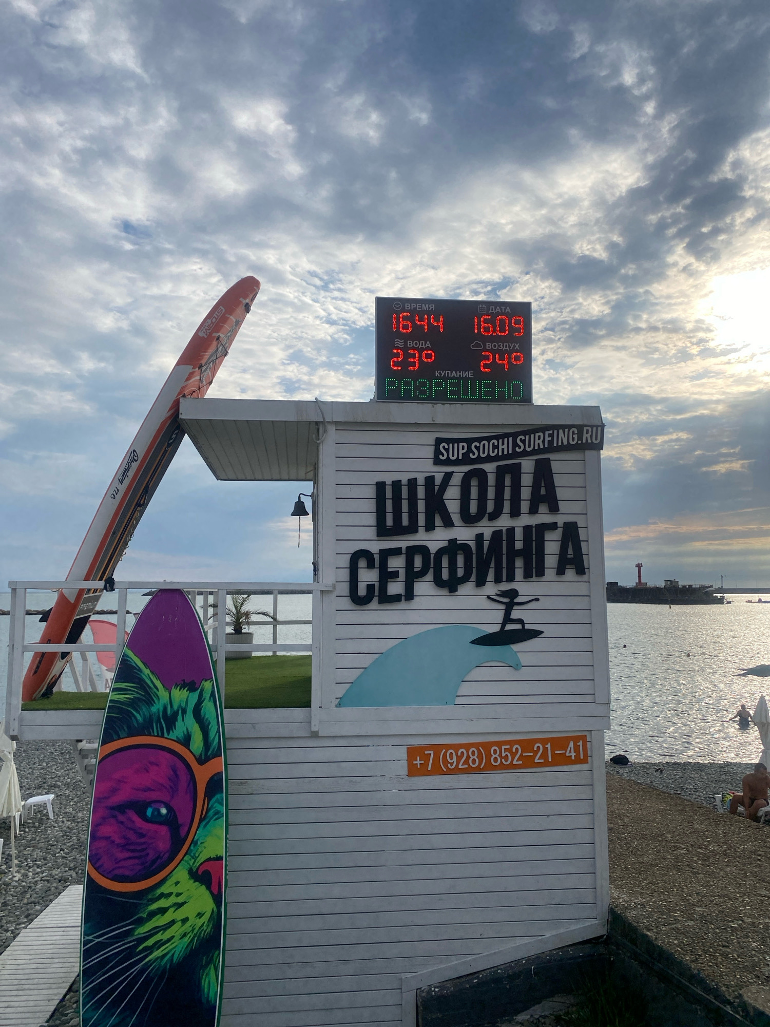 In Sochi, Russia, a surf school building with white horizontal siding is situated on a pebble beach. A digital display board shows the time as 16:44, the date as 16.09, water temperature at 23°, air temperature at 24°, and indicates "SWIMMING IS ALLOWED" in green lettering. Above the display, a white sign reads "SUP.SOCHI.SURFING.RU." The building itself features large black lettering spelling out "ШКОЛА СЕРФИНГА" (Surf School) and a graphic of a surfer on a wave. A bright orange phone number, "+7 (928) 852-21-41," is visible on the side of the building. An orange stand-up paddleboard is propped against the roof of the building, and a surfboard with a colorful cat graphic is standing upright in the foreground. The sea is calm, with a few people visible swimming and relaxing on the beach. In the distance, a red lighthouse stands on a pier. The sky is cloudy with the sun peeking through.