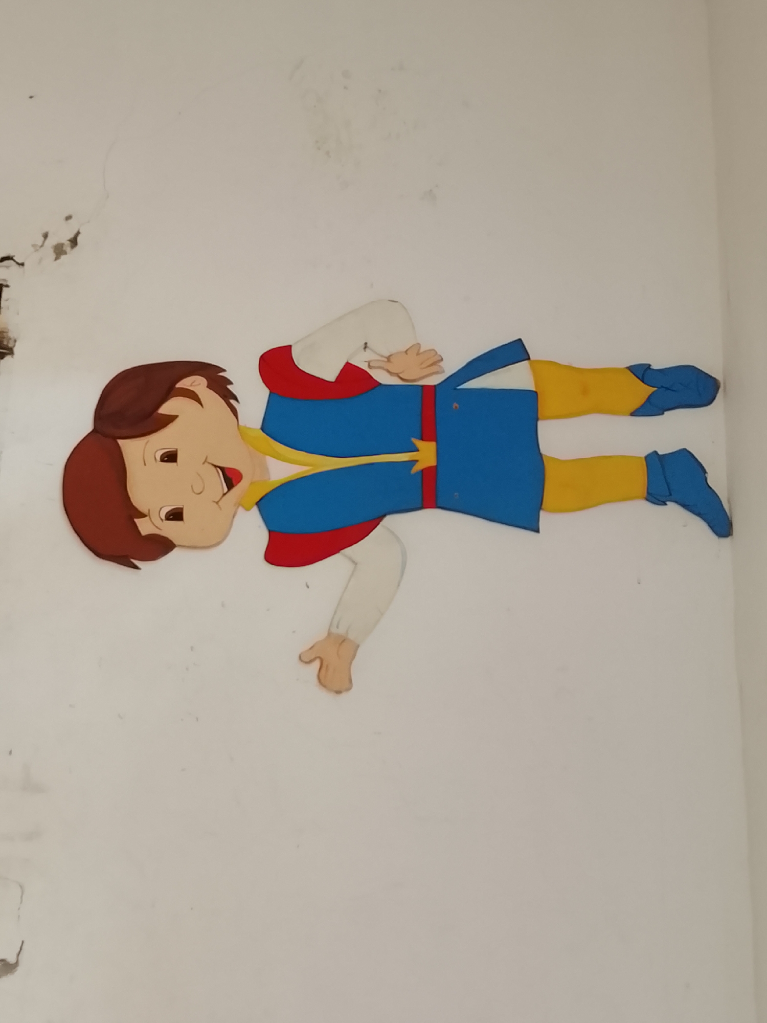 A cartoon cutout of a boy is affixed to a white wall. The cutout is a crude representation of a young boy, dressed in a blue tunic with red trim, yellow leggings, and blue shoes. The boy has brown hair and tan skin. The image quality is low and somewhat blurry. The upper left portion of the wall shows damage/peeling. The location is Altagracia, Venezuela.