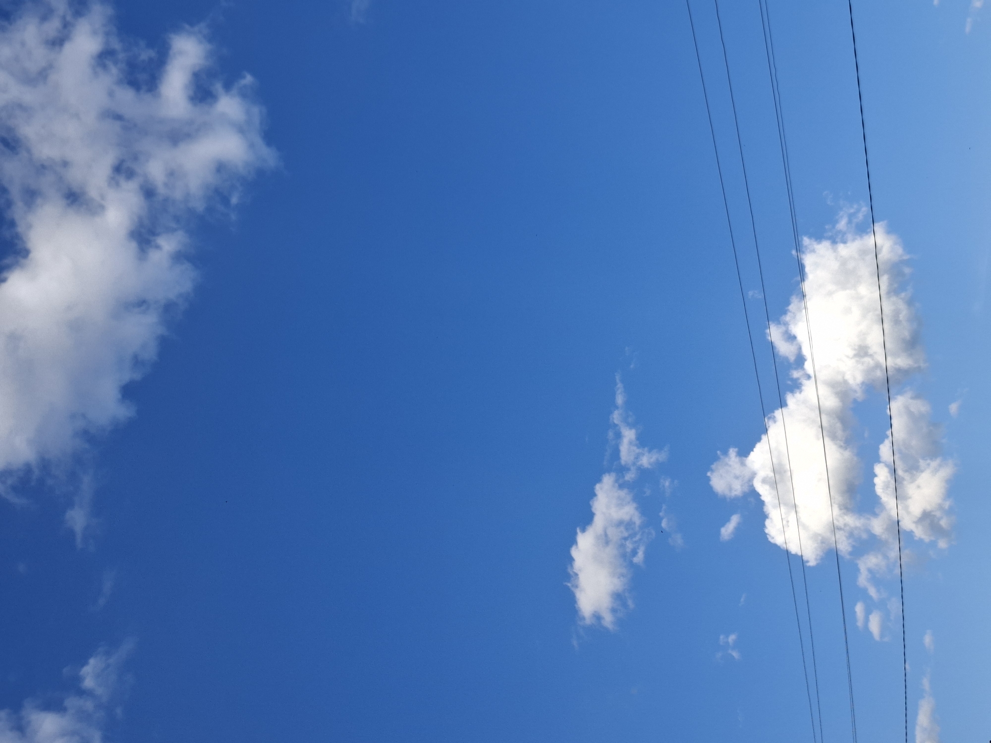 The image is a daytime shot of a bright blue sky with scattered clouds. On the left side of the frame, a puffy white cloud is visible. Toward the right, a larger, denser cloud formation is present, partially obscured by four dark electrical power lines running vertically through the frame. A smaller, wispy cloud is situated below and to the left of the larger cloud formation. The scene lacks any indication of being in Karsha, Russia, with no visible landmarks or cultural elements.