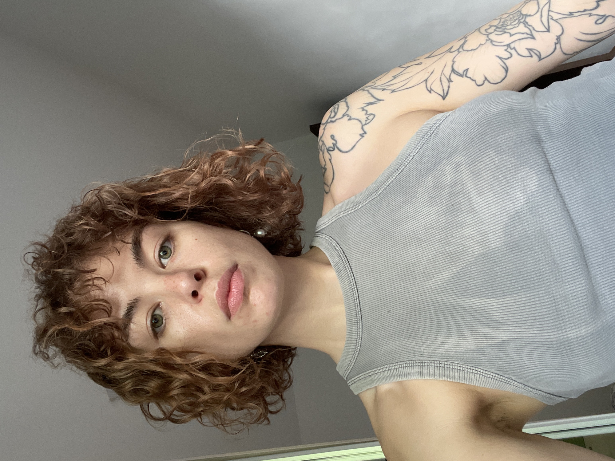 A medium-shot, low-angle view of a young adult female indoors. She has curly reddish-brown hair and green eyes, with fair skin. She is wearing a light gray ribbed tank top. A floral tattoo is visible on her left upper arm and shoulder. The background is a plain, light gray wall. The location is not determinable from the image.