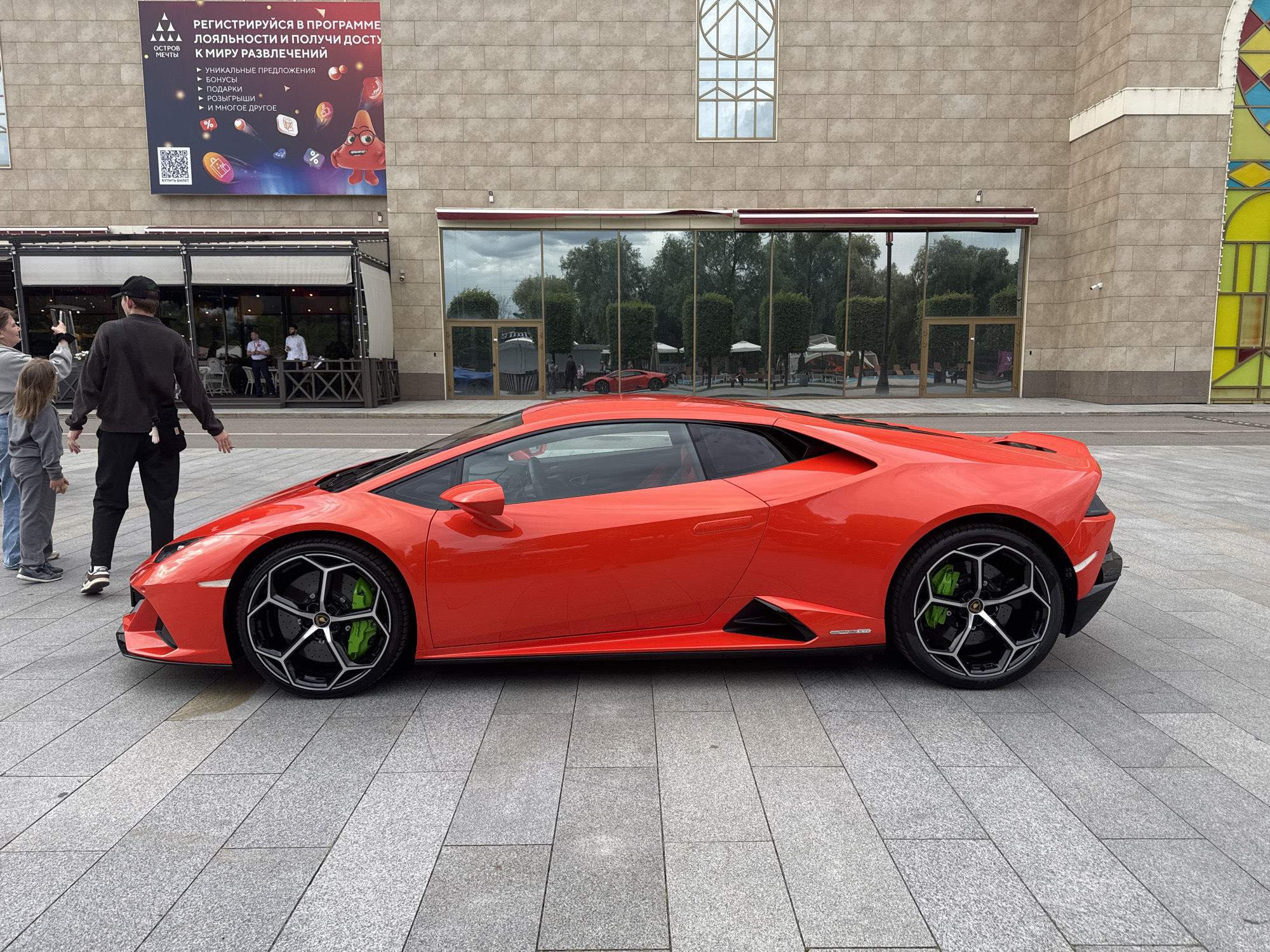 The image shows a red Lamborghini Huracan STO parked on a grey paved surface in front of a building. The building has a large banner with Russian text and a QR code, and large windows reflecting the sky and trees. The car's wheels have black rims and bright green brake calipers. There are a few people standing on the left side of the image, seemingly observing the car. The scene appears to be an outdoor area, possibly a public square or a promenade, located in Moscow, Russia, given the Russian text on the banner.