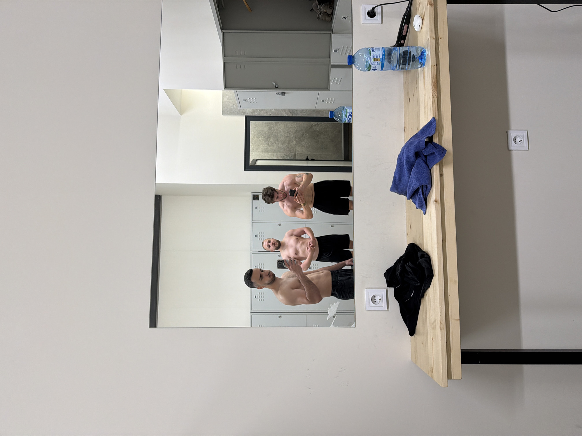 The image appears to be taken inside a changing room, likely in a fitness center or sports facility. The scene is captured through a large mirror. Reflected in the mirror are three shirtless men taking a selfie. The background shows rows of metal lockers. Next to the mirror, there's a light wooden shelf with a water bottle, a blue towel, and a black object (possibly clothing) on it. An electrical outlet is located above the shelf and another one is located on the wall next to the mirror.
The location in Plovdiv, Bulgaria cannot be determined from the image.