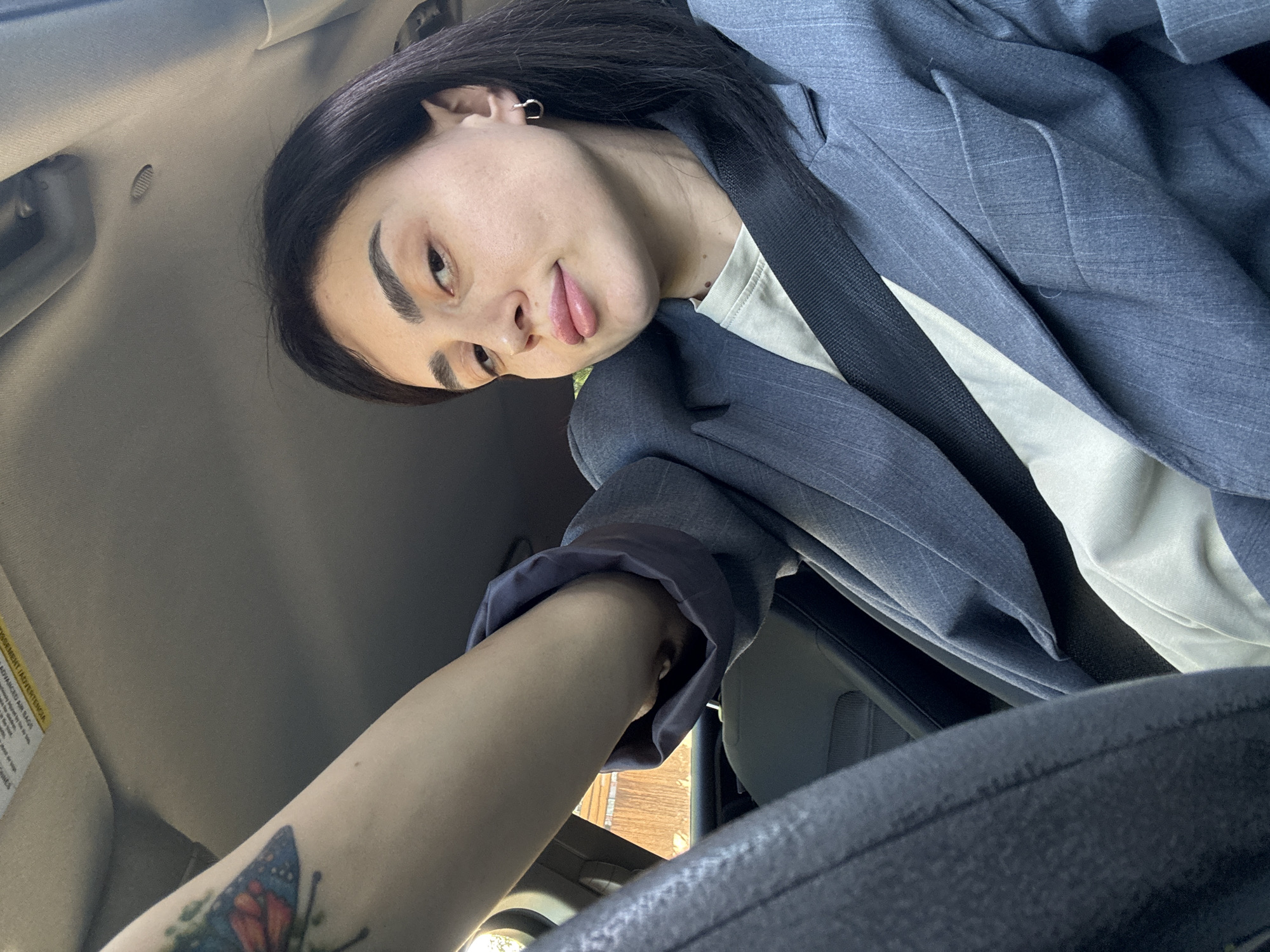 A woman with dark hair and eyebrows, wearing a light blue pinstripe blazer over a white shirt, sits in the driver's seat of a car. The black seatbelt is visible across her chest. Her left arm, adorned with a butterfly tattoo, reaches toward the steering wheel. The car's interior is gray. The view through the car window shows a sunny outdoor setting, potentially in Tbilisi, Georgia, as a building with reddish-brown bricks is visible.