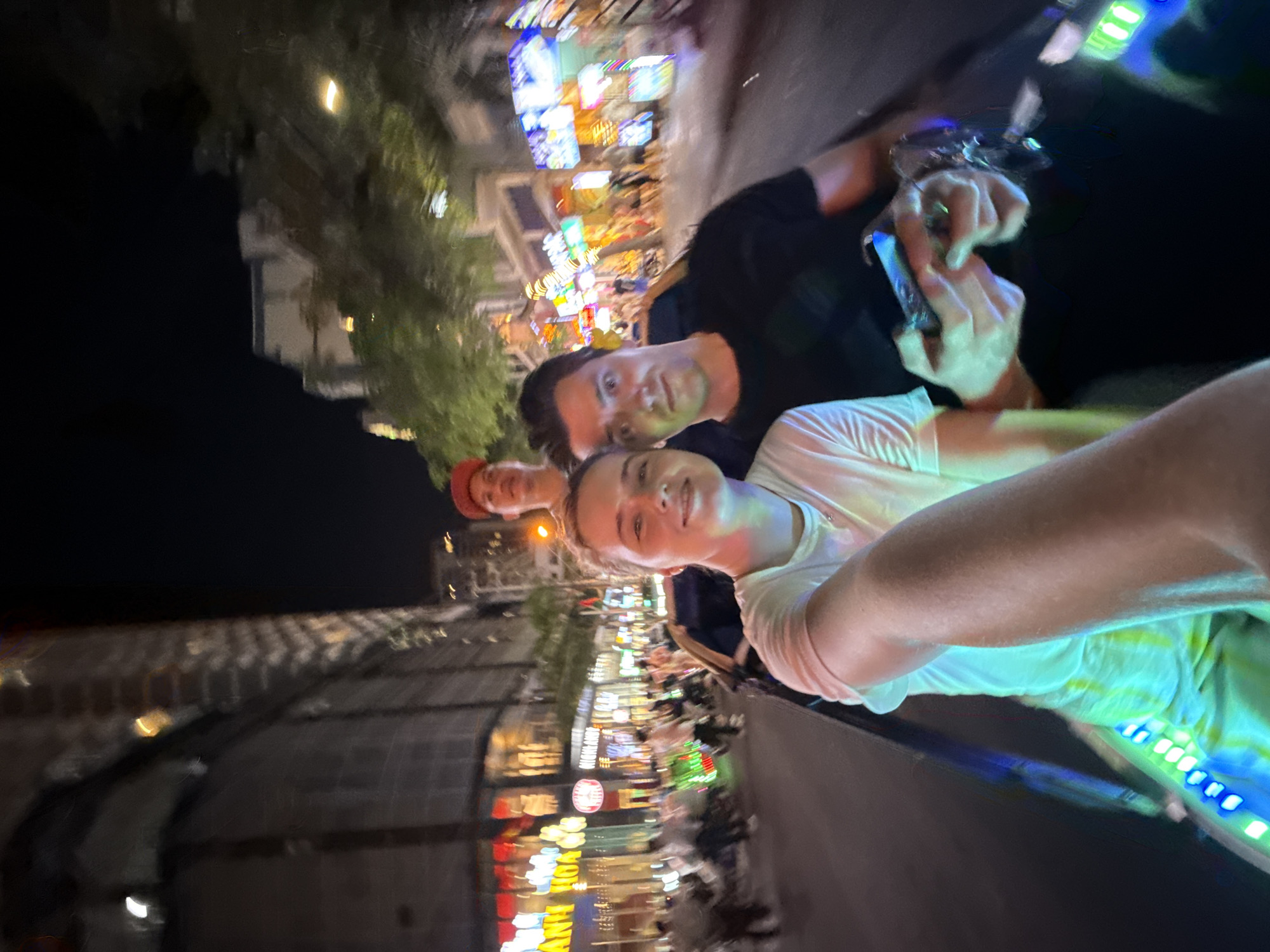 This sideways-oriented night photograph captures three individuals in a lively urban setting, likely Nha Trang, Vietnam. In the foreground, positioned on the left side of the frame, a person with light skin wearing a white t-shirt smiles faintly towards the camera. Directly behind them and slightly to the right, another person with light skin, sporting a black t-shirt, also looks towards the camera while holding what appears to be a smartphone or small device with both hands. A third person wearing a red cap is partially visible on the far right, behind the second individual.
The background, appearing blurry due to motion or low light, reveals a bustling street scene lined with multi-story buildings. Numerous vibrant and colorful illuminated signs and storefronts, typical of a commercial district at night, stretch into the distance. Bright streaks of light and reflections indicate a dynamic environment. On one prominent yellow sign, the Vietnamese text "ANH HOA" is discernible. The immediate foreground on the right side features glowing blue and green LED strips, suggesting an active, modern street or perhaps a feature of a vehicle they might be on. The overall mood is casual and pleasant, indicative of a night out or evening exploration.