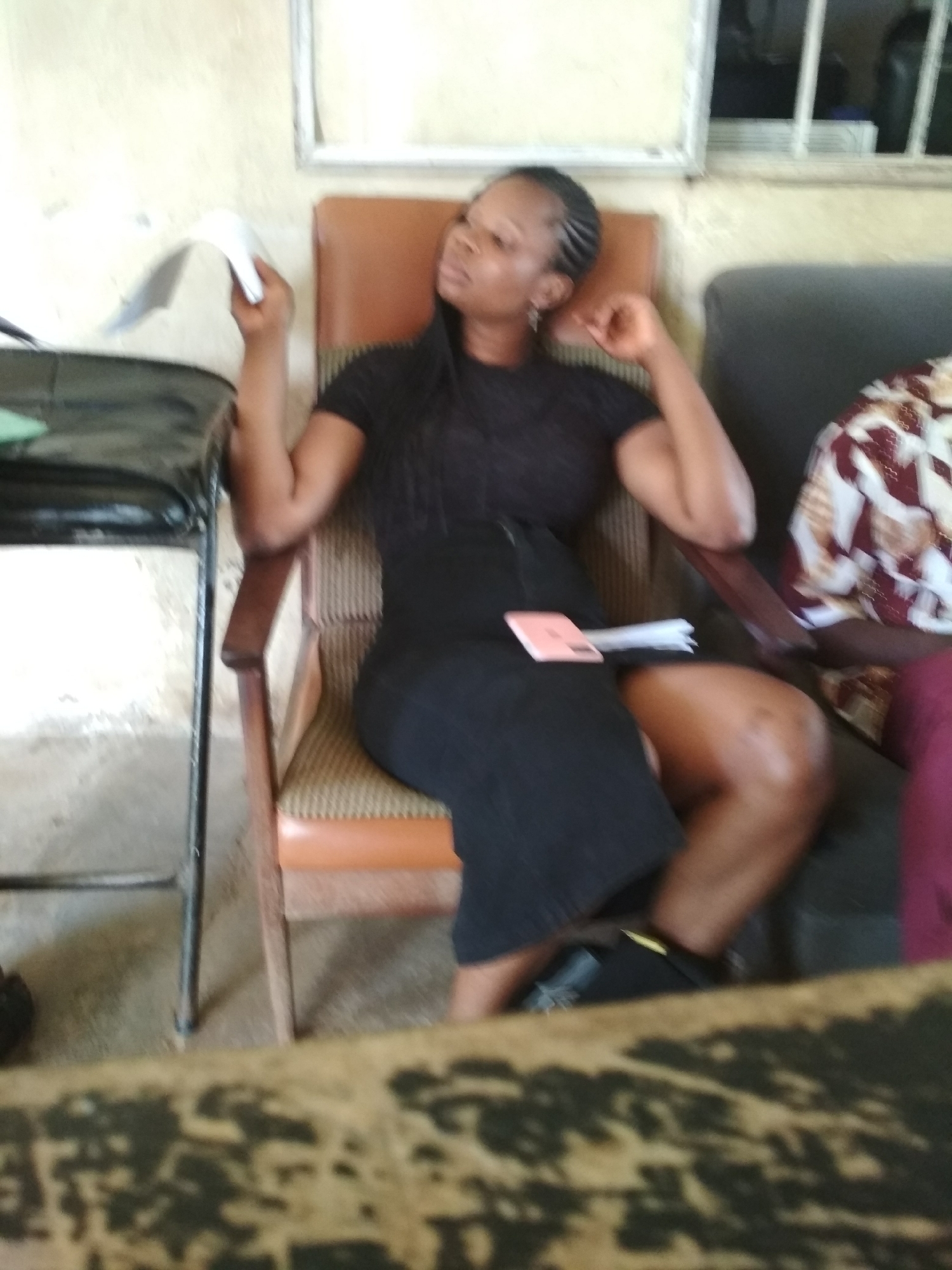 A woman with braided hair is seated in a chair, holding a white paper. She is wearing a black dress and is positioned in a room with light-colored walls. To her right, another person is partially visible wearing a patterned garment. In the foreground, a dark surface with a textured pattern is present. The overall lighting is dim. The setting appears to be an indoor space in Bisichi, Nigeria.