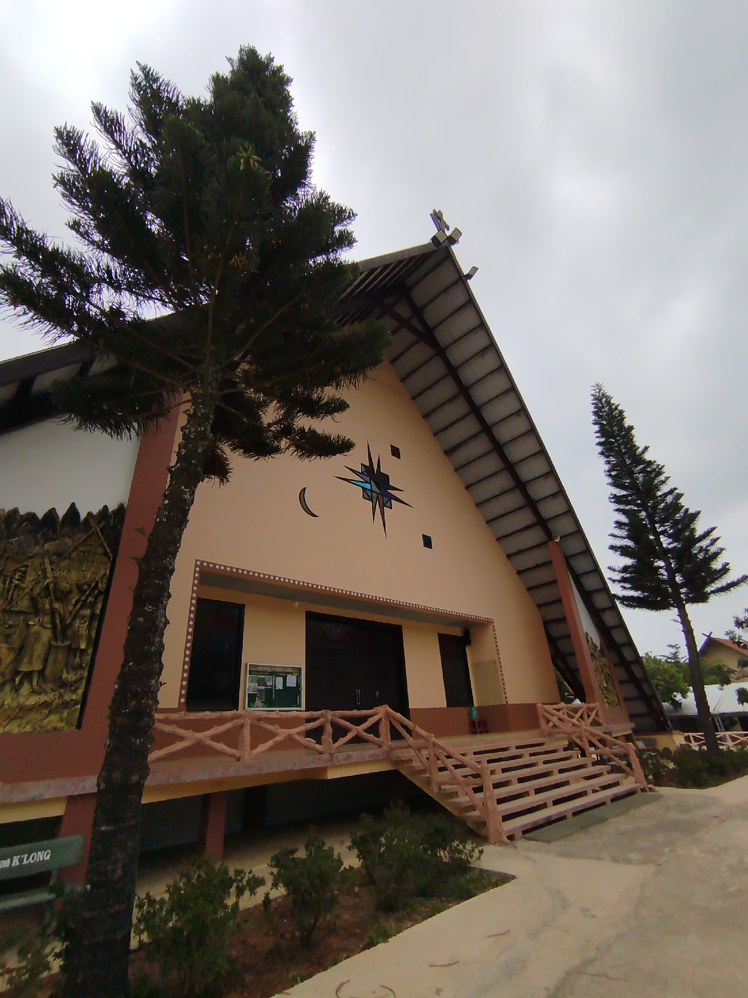 The image depicts a church in K'Long, Vietnam. The building features a peaked roof with a cross at the apex. A crescent moon and star design adorn the facade, along with square insets. The entrance is accessed via concrete steps and a low, decorative railing. Relief sculptures appear on the side walls of the building. Two coniferous trees are located near the church, one prominent on the left and another on the right. A concrete pathway leads to the church entrance. A bench and vegetation are visible in the foreground. The sky is overcast.