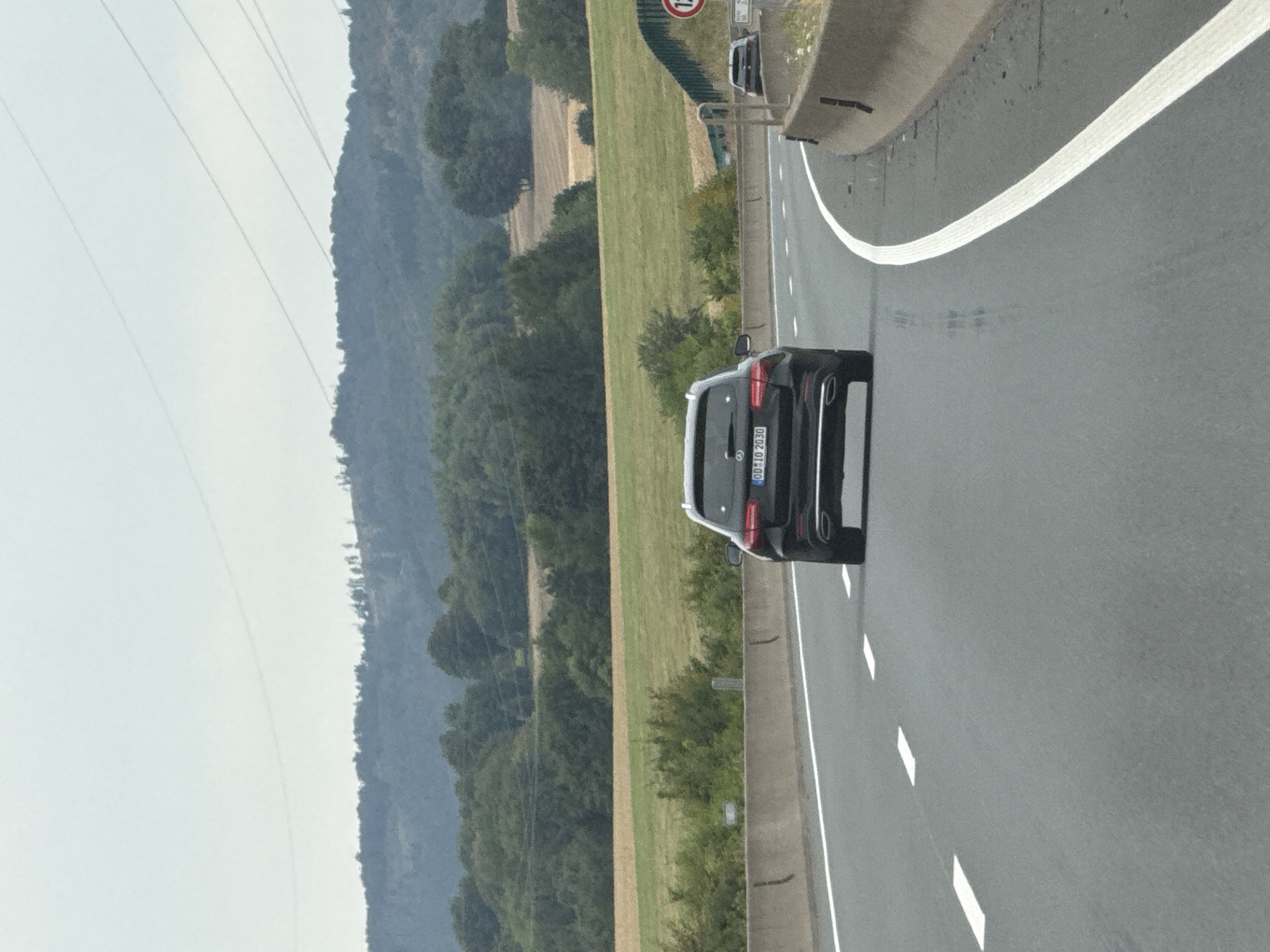 An aerial view from a vehicle on a highway shows a black Mercedes-Benz SUV driving in the right lane. The car has a license plate that reads "OD IO 2030". The highway has white lane markings and concrete barriers. In the background, there are rolling hills covered with trees, fields, and power lines stretching across the sky. A speed limit sign indicating 120 kilometers per hour is visible on the right side of the road. The weather appears to be overcast. The image does not provide specific details about the city of Bad Lauterberg im Harz, Germany, beyond the highway context.