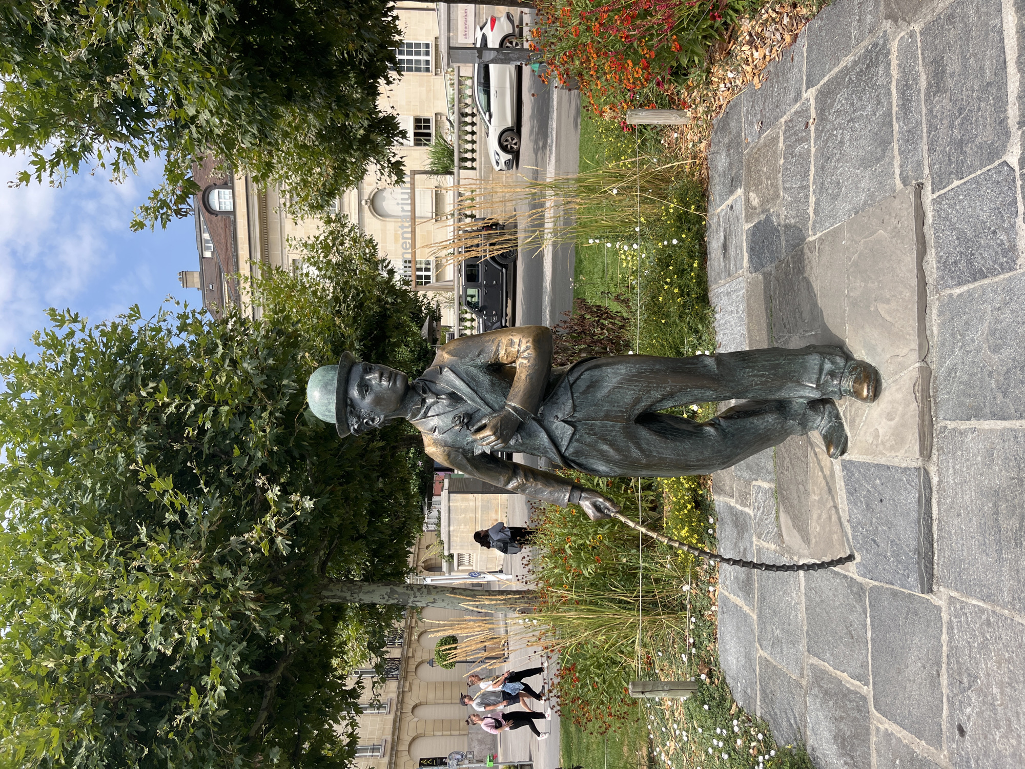 The image shows a bronze statue of Charlie Chaplin in Vevey, Switzerland. The statue is located in a public square, with a paved walkway in the foreground and a garden with flowers and tall grasses behind it. In the background, there is a street with cars and buildings.
The statue depicts Chaplin in his iconic Tramp persona, wearing a bowler hat, suit, and holding a cane. He is standing in a relaxed pose, looking off to the side.
The location of the statue is in front of the Alimentarium, a food museum, in Vevey, Switzerland. The square is a popular spot for tourists and locals alike, and the statue is a beloved landmark.
The image is taken from a slightly low angle, looking up at the statue. The lighting is bright and sunny, with some shadows cast by the trees. The sky is blue with a few clouds.
Several people are visible in the background, walking on the sidewalk and across the street. A dark Jeep and a white car are on the street.
The overall scene is one of a pleasant day in a charming Swiss town, with the statue of Charlie Chaplin serving as a focal point.