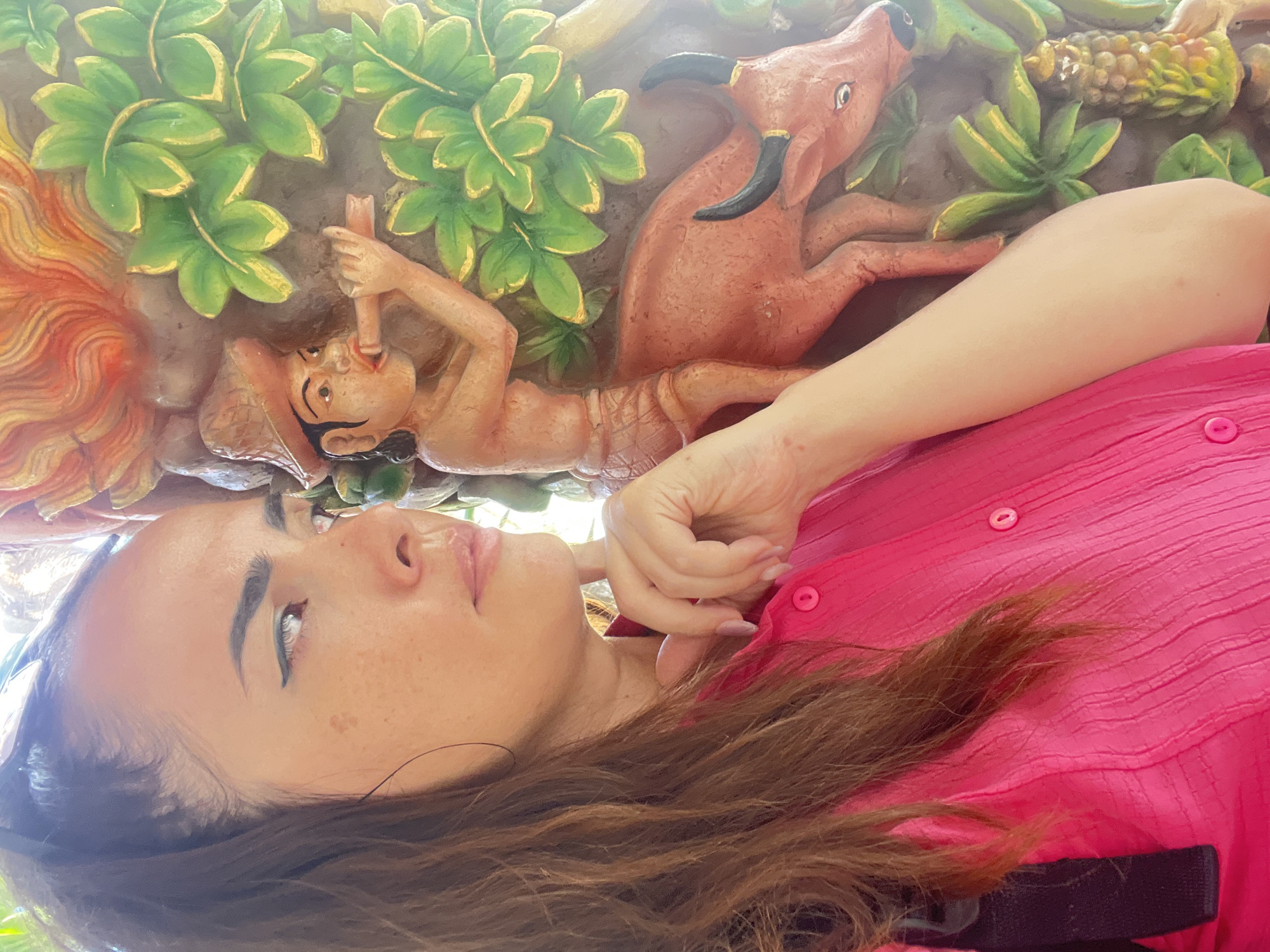 A woman with dark hair and a bright pink shirt poses for a portrait, her head tilted to the right. She is positioned in front of a colorful relief carving that depicts a man playing a flute atop a brown bull. Green leaves and a fiery orange element frame the scene. The woman's left hand is raised, with her fingers touching her chin. The carving appears to be a decorative element on a building in Kuta Selatan, Indonesia.