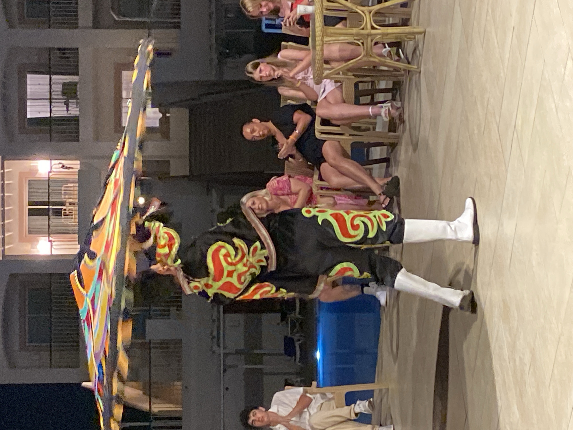This is a medium shot of a Tanoura dancer performing in front of an audience in Sharm El Sheikh, Egypt. The dancer, wearing a traditional black costume with red and green embellishments and white boots, is spinning a large, colorful skirted disc above his head. In the background, several people are seated, observing the performance. To the left of the dancer, a young man sits in a white shirt and beige pants. Behind him, a blue swimming pool with lounge chairs is visible. To the right of the dancer, a group of women and men are seated on chairs, some with drinks on a table. The scene appears to be set at an outdoor venue, possibly a hotel courtyard or restaurant, at dusk or night. The building in the background has multiple balconies and lit windows.