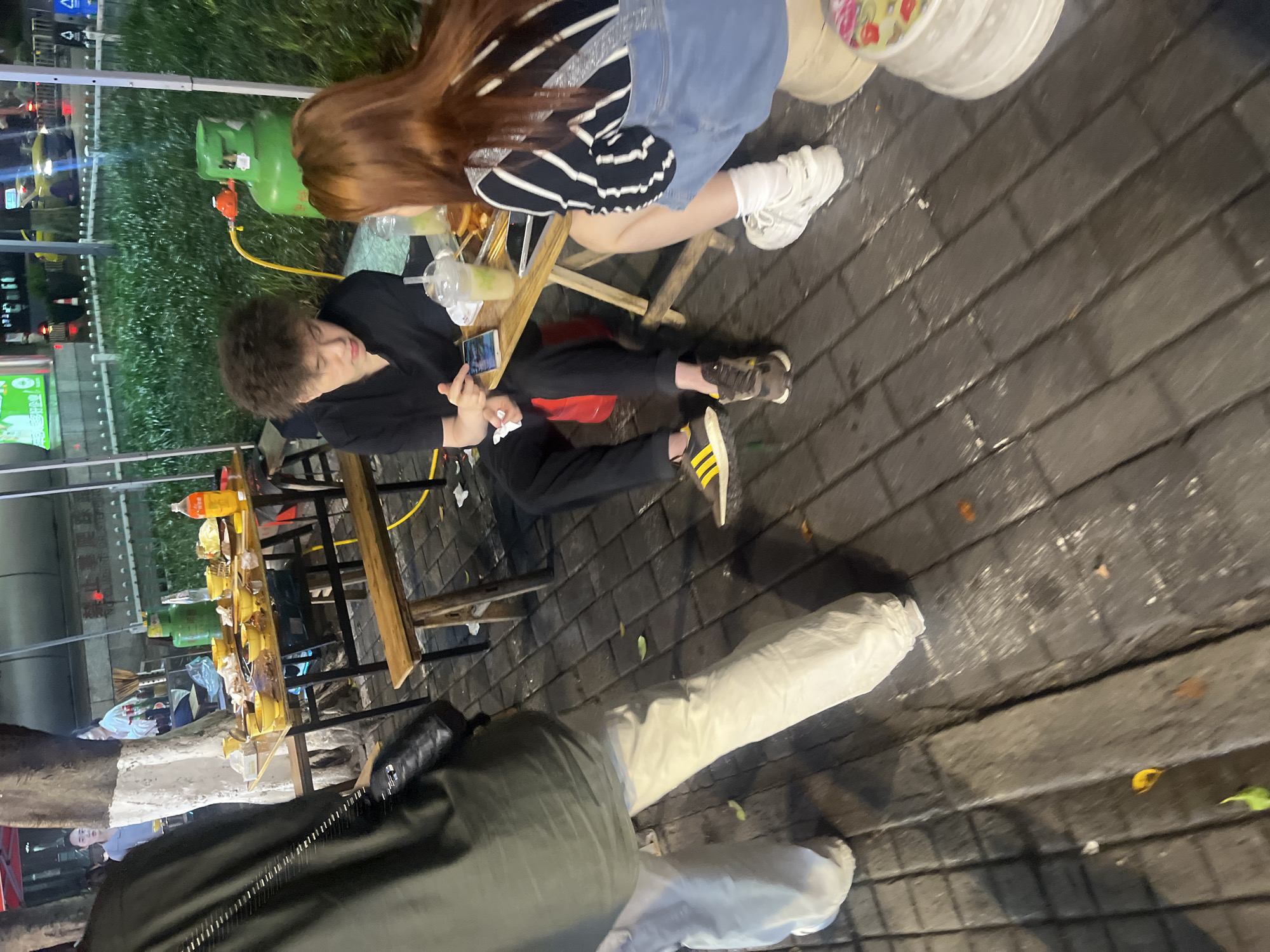 The image depicts an outdoor dining scene at night in Yuzhong District, China. Two young adults, a male with dark, curly hair wearing a black t-shirt and black pants, and a female with reddish-brown hair wearing a black and white striped shirt and a denim skirt, are seated at a small wooden table. The male is holding a smartphone and appears to be showing it to the female. There are drinks and food on the table. The ground is paved with dark brick. In the background, there is a large green gas tank, a tree, and other diners. A person's legs are visible in the foreground on the left, wearing light-colored pants.