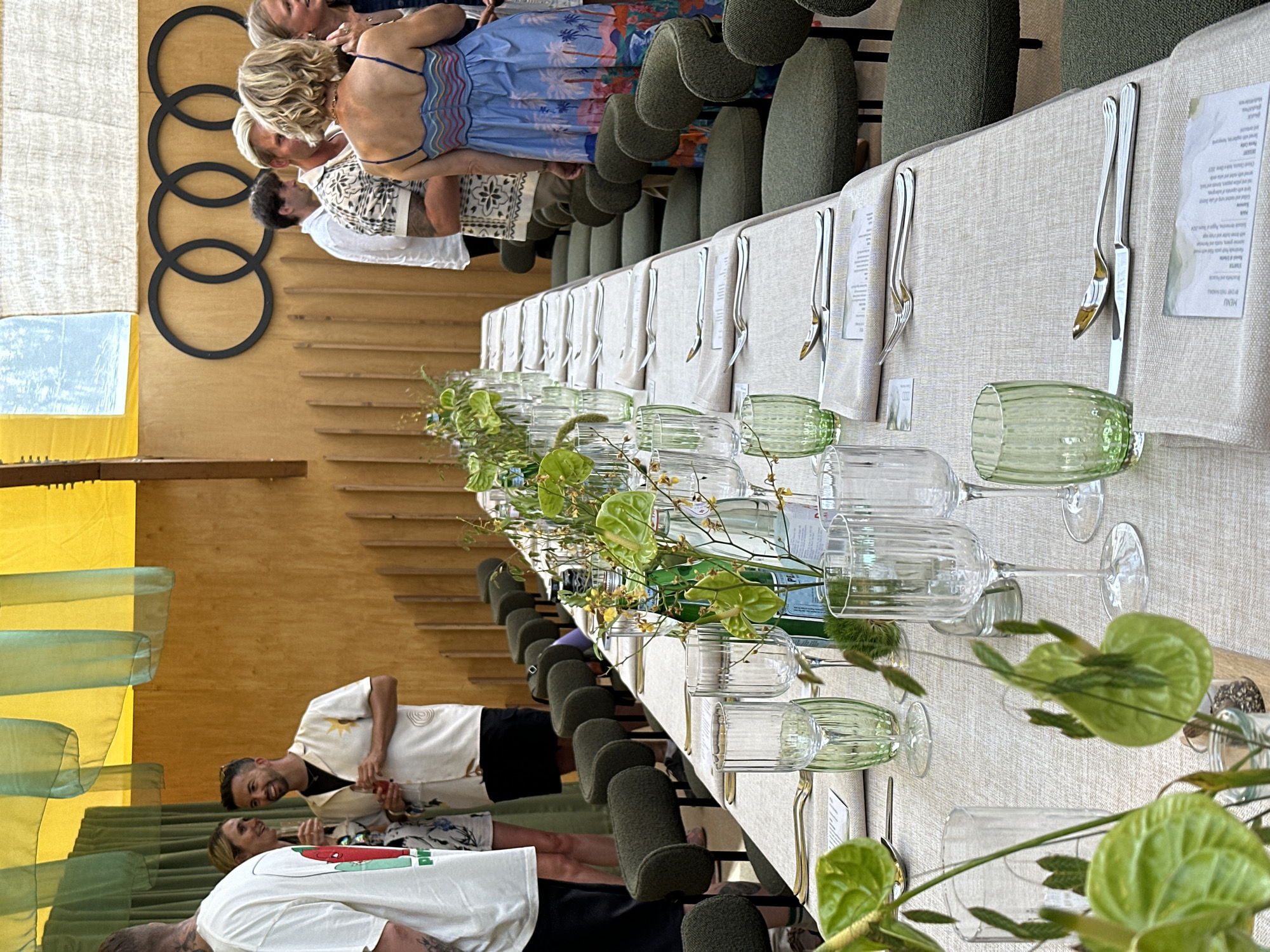 An indoor event space is depicted, with a long, set dining table as the central focus. The table is covered with a light-colored linen cloth and is adorned with green glassware, clear wine glasses, silverware, and floral arrangements featuring green anthurium flowers. Several bottles of water are also placed along the table. Green upholstered chairs are arranged on either side of the table. In the background, a wooden wall features a large Audi logo, composed of four interlocking rings. Several individuals are standing and conversing around the table. One man wears a white shirt with a sun graphic, another a black t-shirt, and a woman is seen in a blue dress with a palm tree print. The ceiling is decorated with yellow and green fabric drapes.