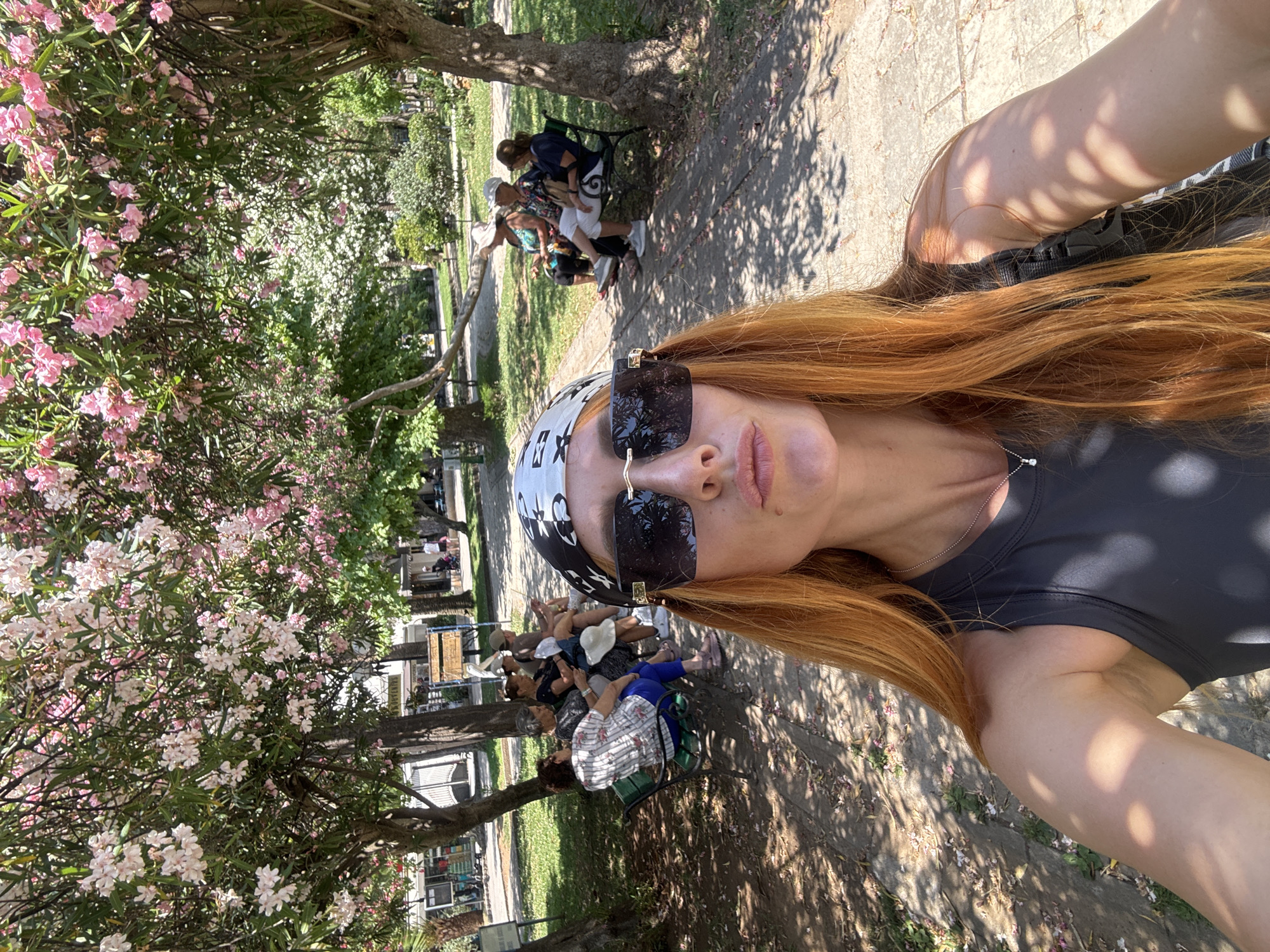 In Budva, Montenegro, a woman with long red hair, wearing a black bandana, large black sunglasses, and a dark grey tank top, takes a selfie in a park. She is in the foreground, with her face angled towards the camera, puckering her lips. Behind her, several elderly women are seated on green park benches, some reading and others chatting. The park features a paved walkway, trees with blooming pink and white flowers, and lush green grass. Sunlight filters through the trees, creating dappled shadows on the ground. The overall scene suggests a pleasant day in a public outdoor space.