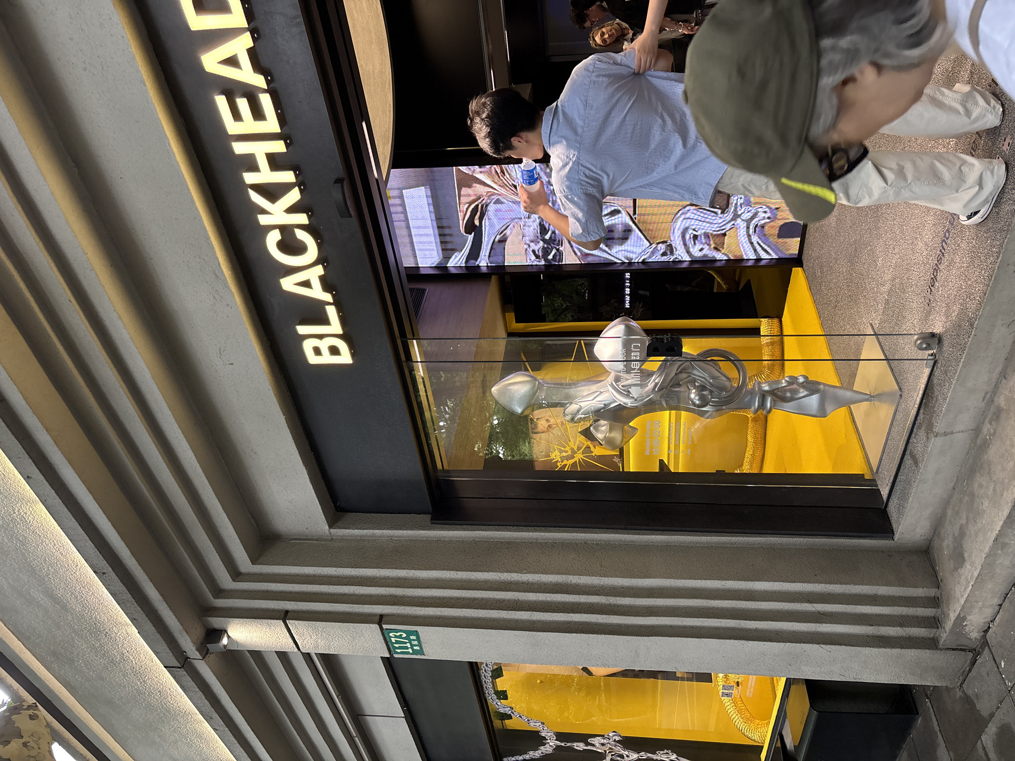 In Changning District, China, the facade of a store with a prominent "BLACKHEAD" sign features a large glass window displaying a bright yellow interior with what appears to be a sculpture. In front of the store, a man in a striped shirt drinks from a bottle while looking at a digital screen displaying abstract imagery. A person wearing a green cap stands in the foreground, looking down at the ground. Another person with dark hair is partially visible to the right. The store is illuminated by overhead lighting and has a modern architectural design with vertical grooved elements on the exterior. A street number "1173" is visible on a green sign.