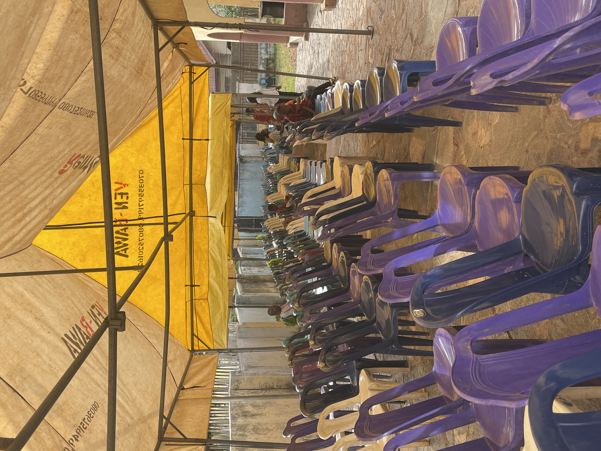 The image shows rows of stacked plastic chairs under a tent, likely set up for an event in Akwanga, Nigeria. The tent is supported by metal poles and has a yellow and beige canopy with writing on it. Some people are sitting in the chairs toward the end of the row. The ground is stone or concrete.