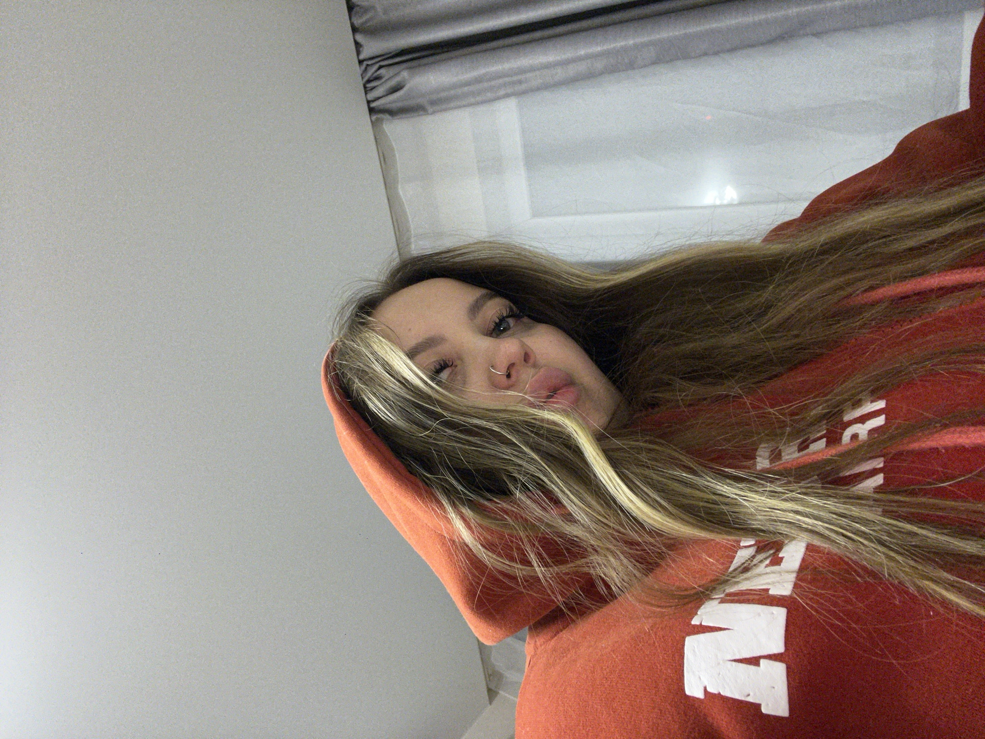 A young woman with long, blonde-streaked hair is wearing an orange hoodie with white lettering. She has a nose ring and is looking at the camera. The background includes a grey wall and a window with grey blinds. There is no location information, so this description cannot include the city of Krasnodar, Russia.